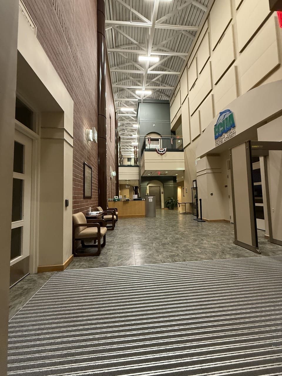 A long, tall corridor with a high ceiling, brick and white walls, chairs on the left, and a central reception desk.