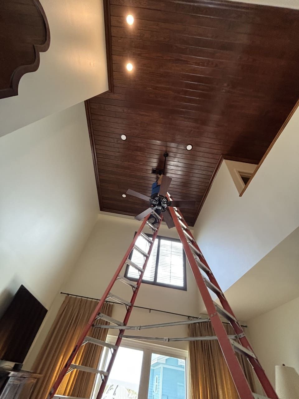 A person on a tall ladder reaches up to install a ceiling fan on a high, dark wood-paneled ceiling in a bright room.