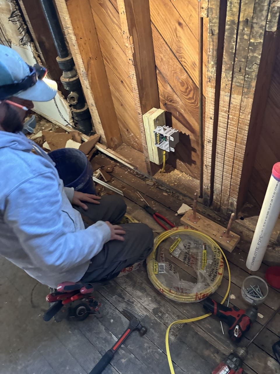 A person kneeling in a construction zone, installing an electrical box onto a wooden wall stud near tools and wiring.