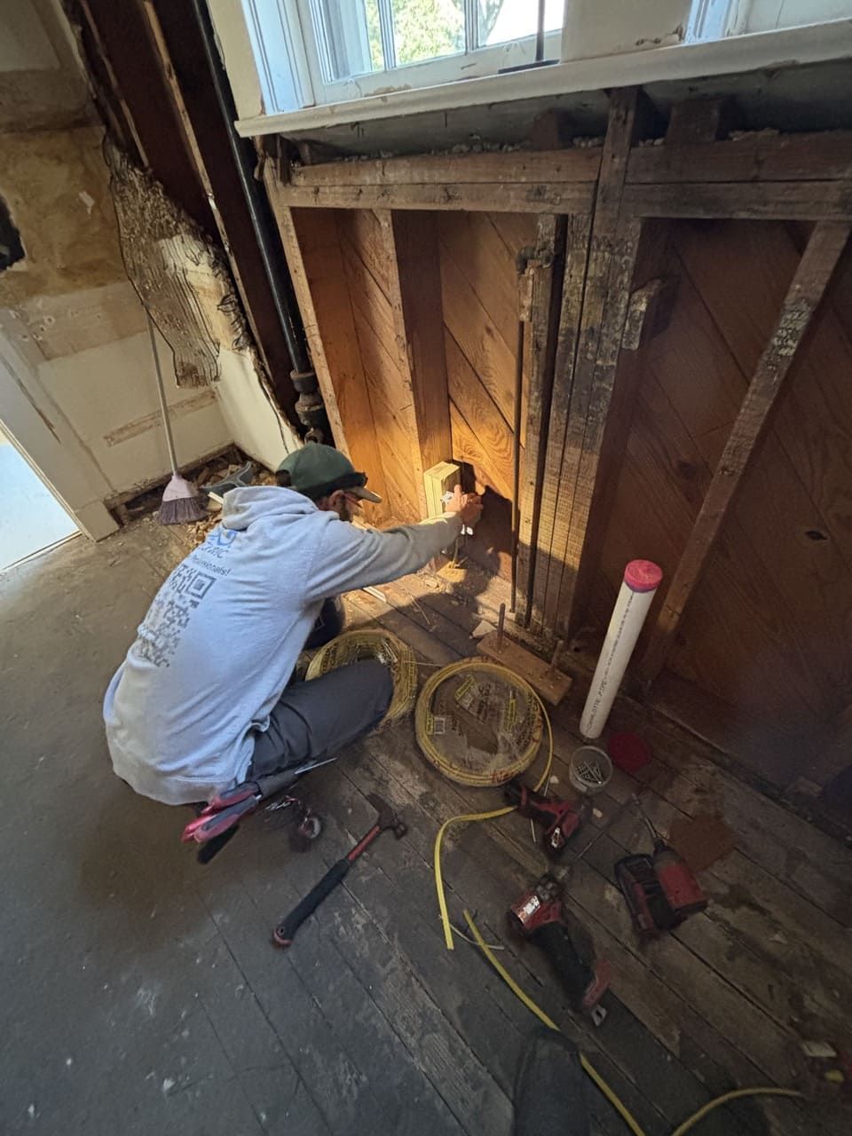 A person in a gray hoodie works inside an open wall cavity under a window, with tools and construction debris nearby.