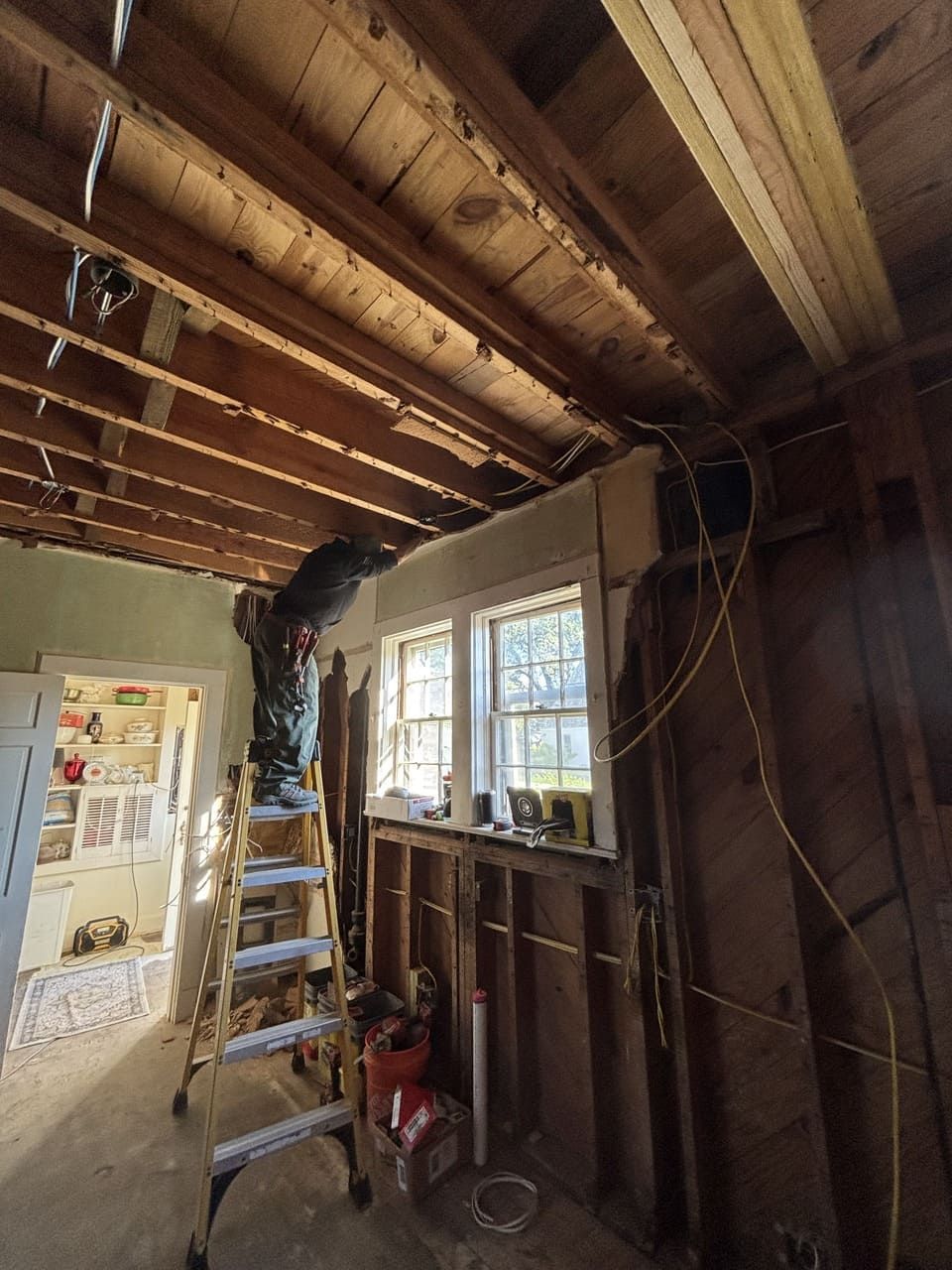 A person standing on a ladder works on exposed wooden ceiling joists in a room undergoing interior renovation.