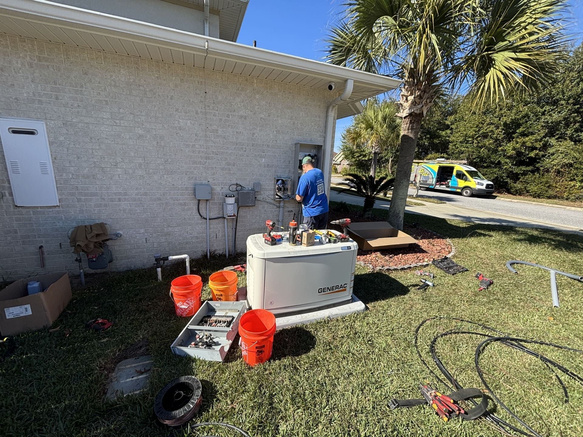 A technician installs a standby generator outside a house near a parked service van and landscaping tools.