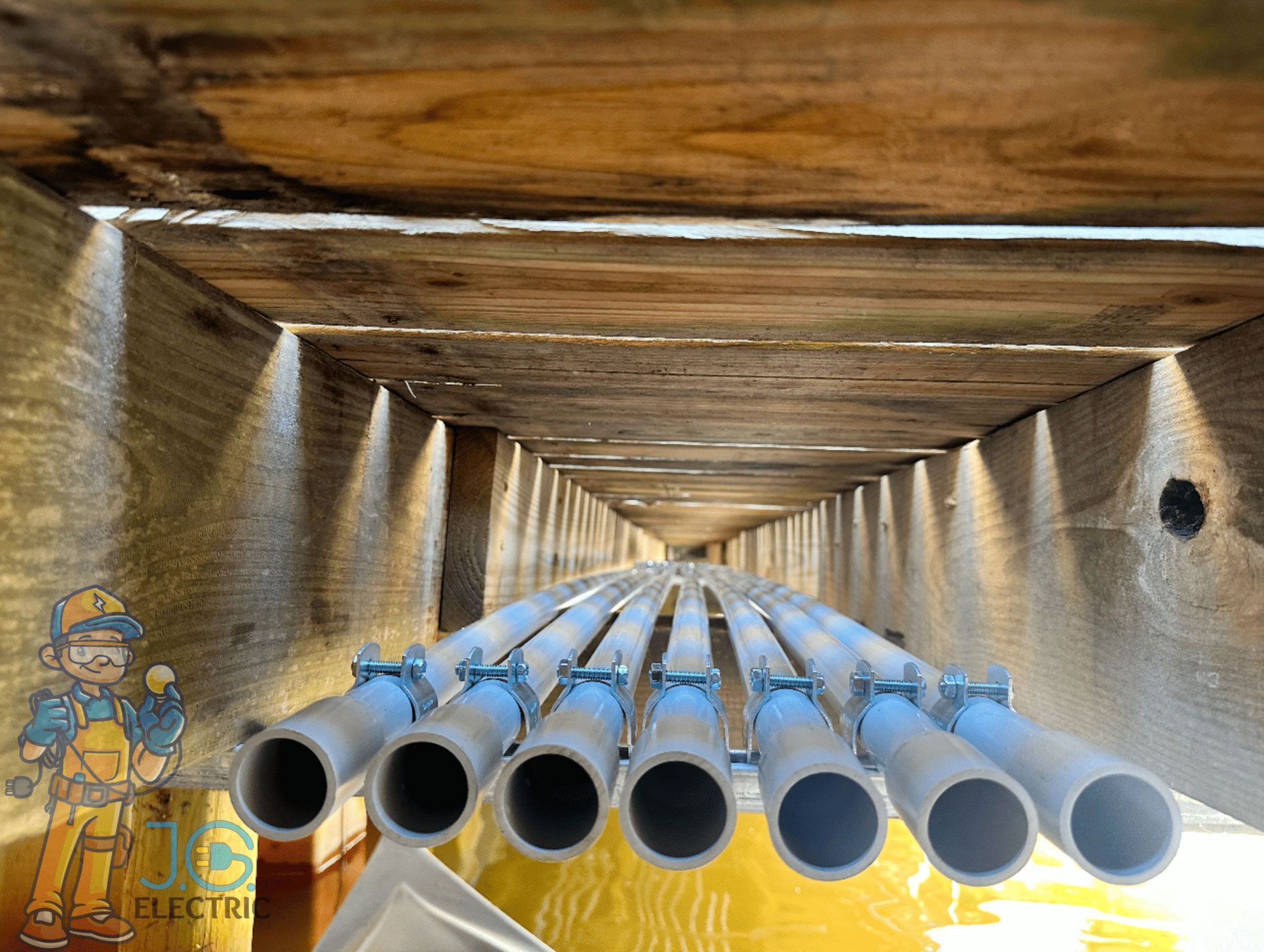 Multiple gray conduits mounted under a wooden structure, with a cartoon electrician graphic in the corner.