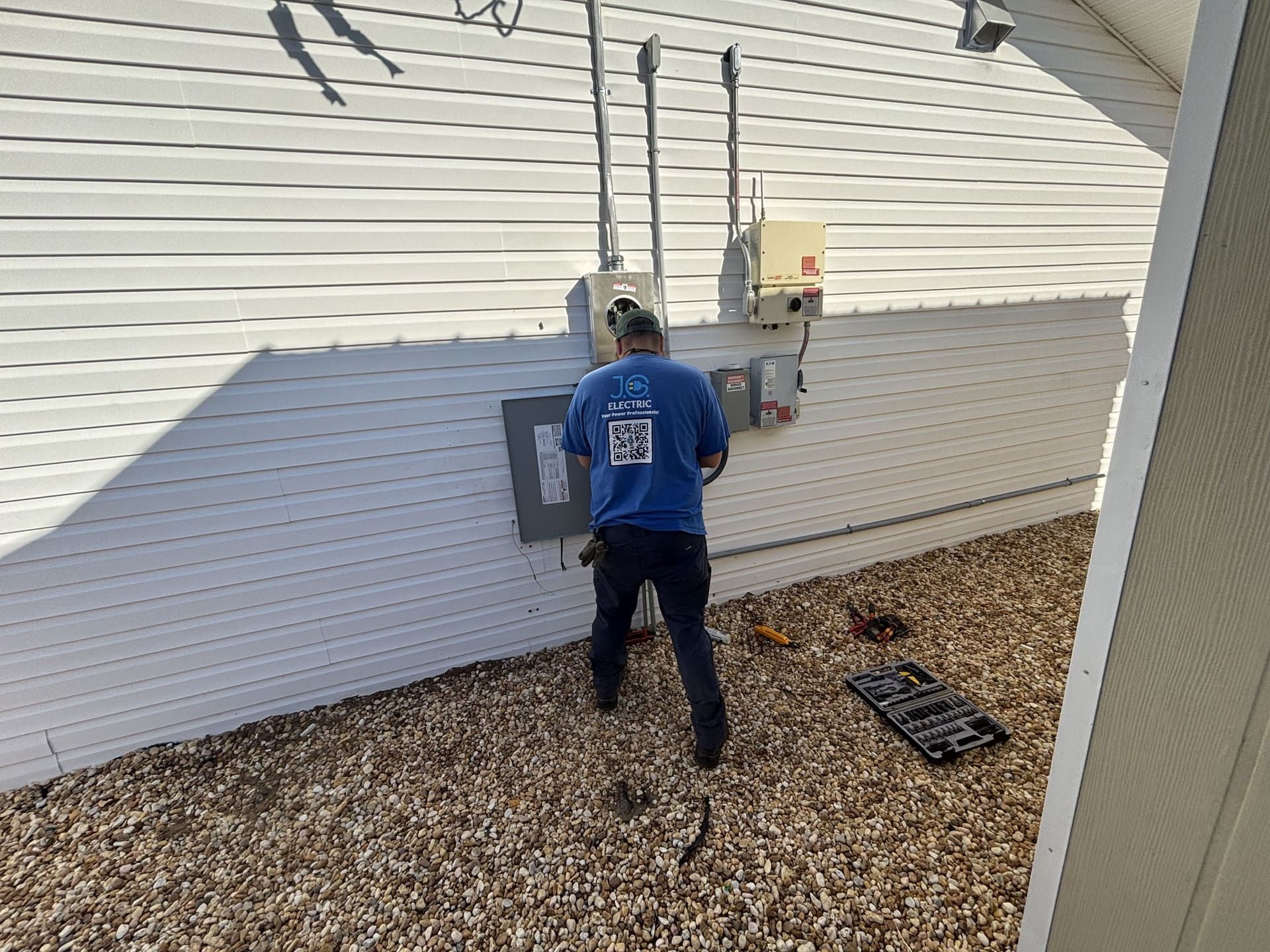An electrician in a blue shirt works on electrical equipment mounted to the exterior of a white-sided building.