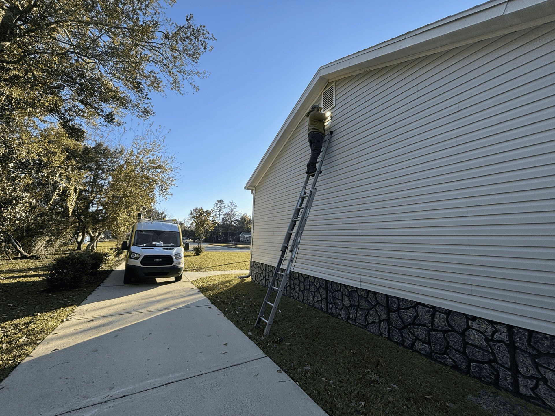 A worker on an extension ladder works on a white house exterior near a driveway with a parked van in the background.