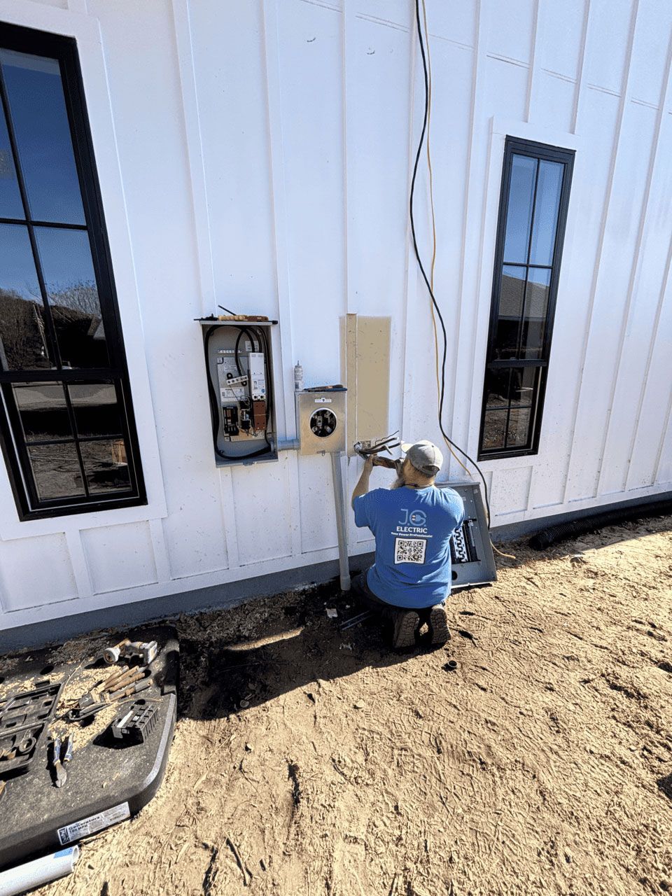 An electrician in a blue shirt kneels outside, installing electrical equipment on a white board-and-batten exterior wall.