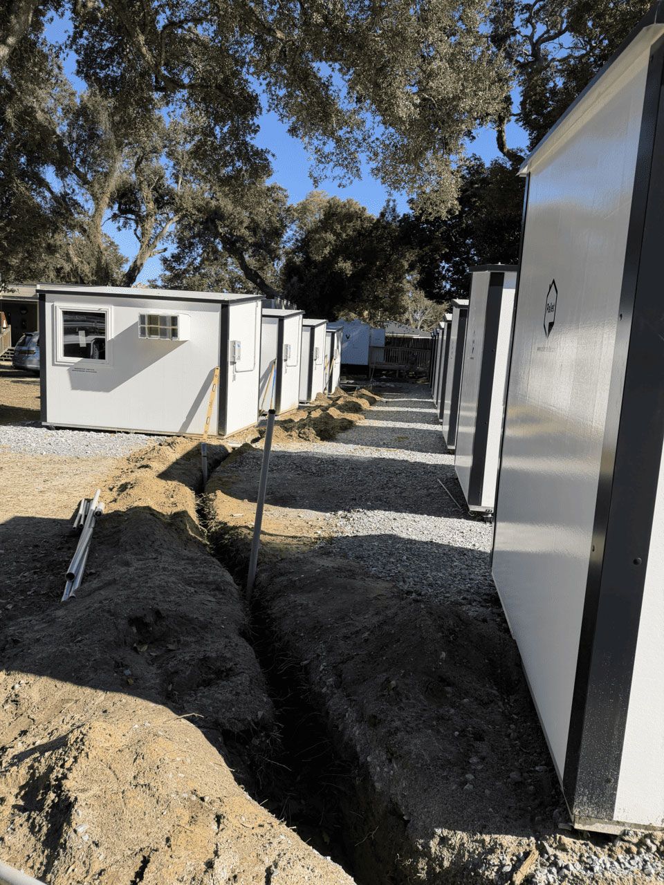 A row of white modular cabins under trees, with an open trench in the foreground on a gravel site.