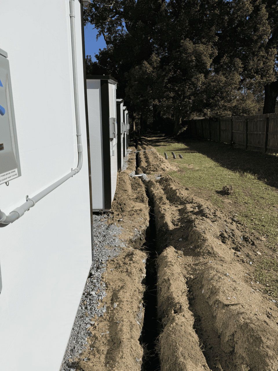 A trench dug in the ground runs parallel to a row of white outdoor industrial cabinets on a grassy lot.