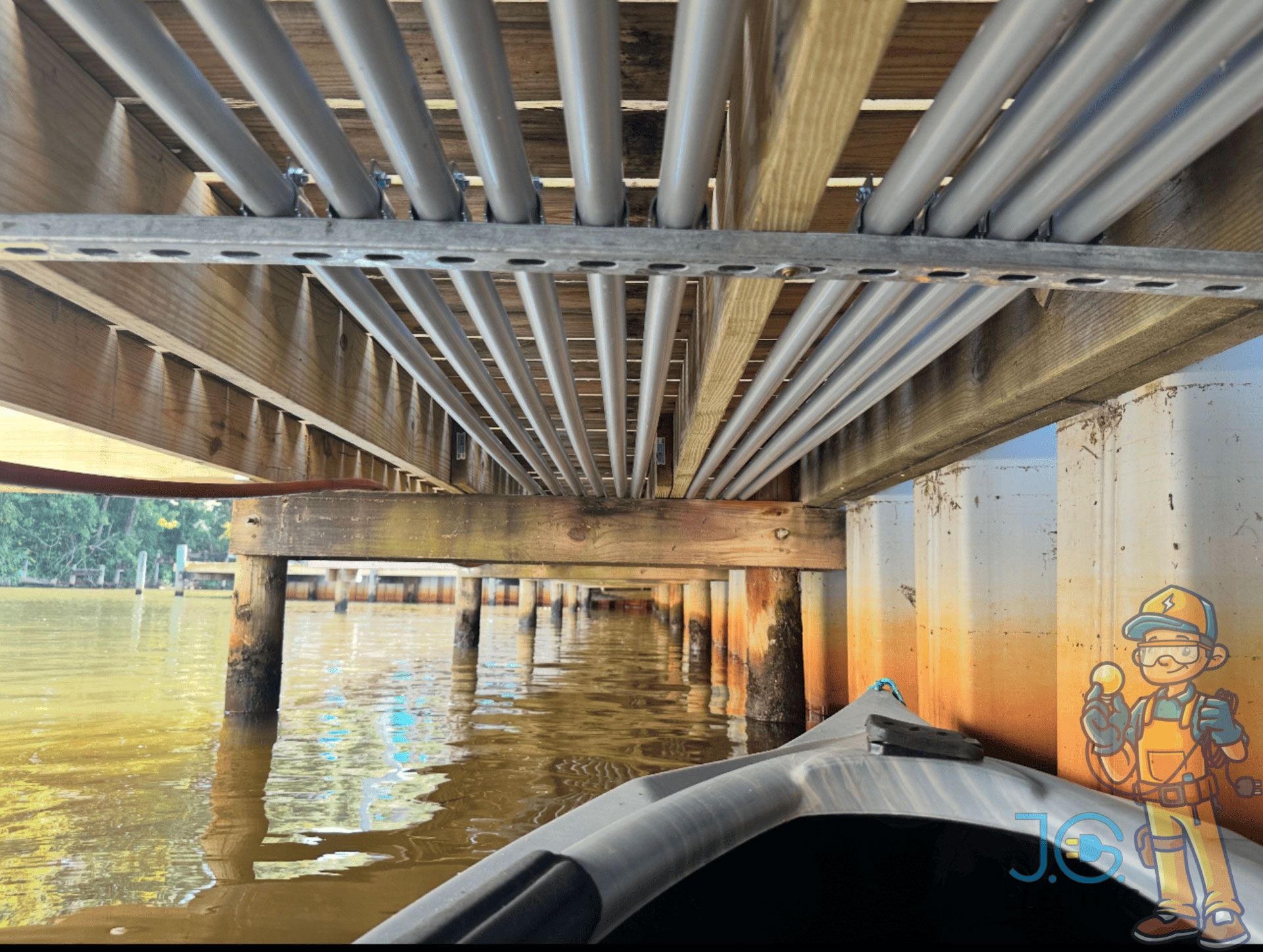View from a kayak underneath a wooden pier showing multiple parallel gray electrical conduits attached to the beams.