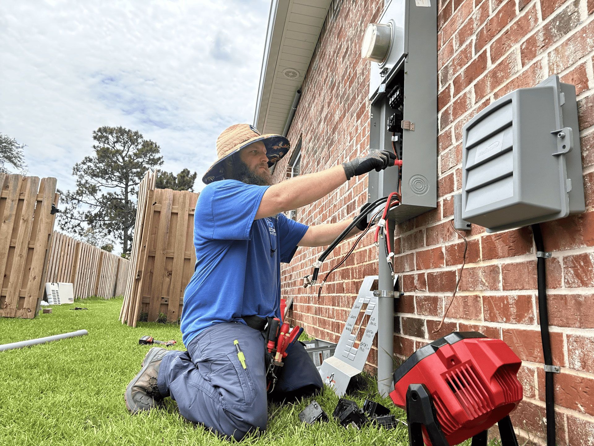 A person in a straw hat and blue shirt kneeling on grass while working on an open electrical box attached to a brick wall.