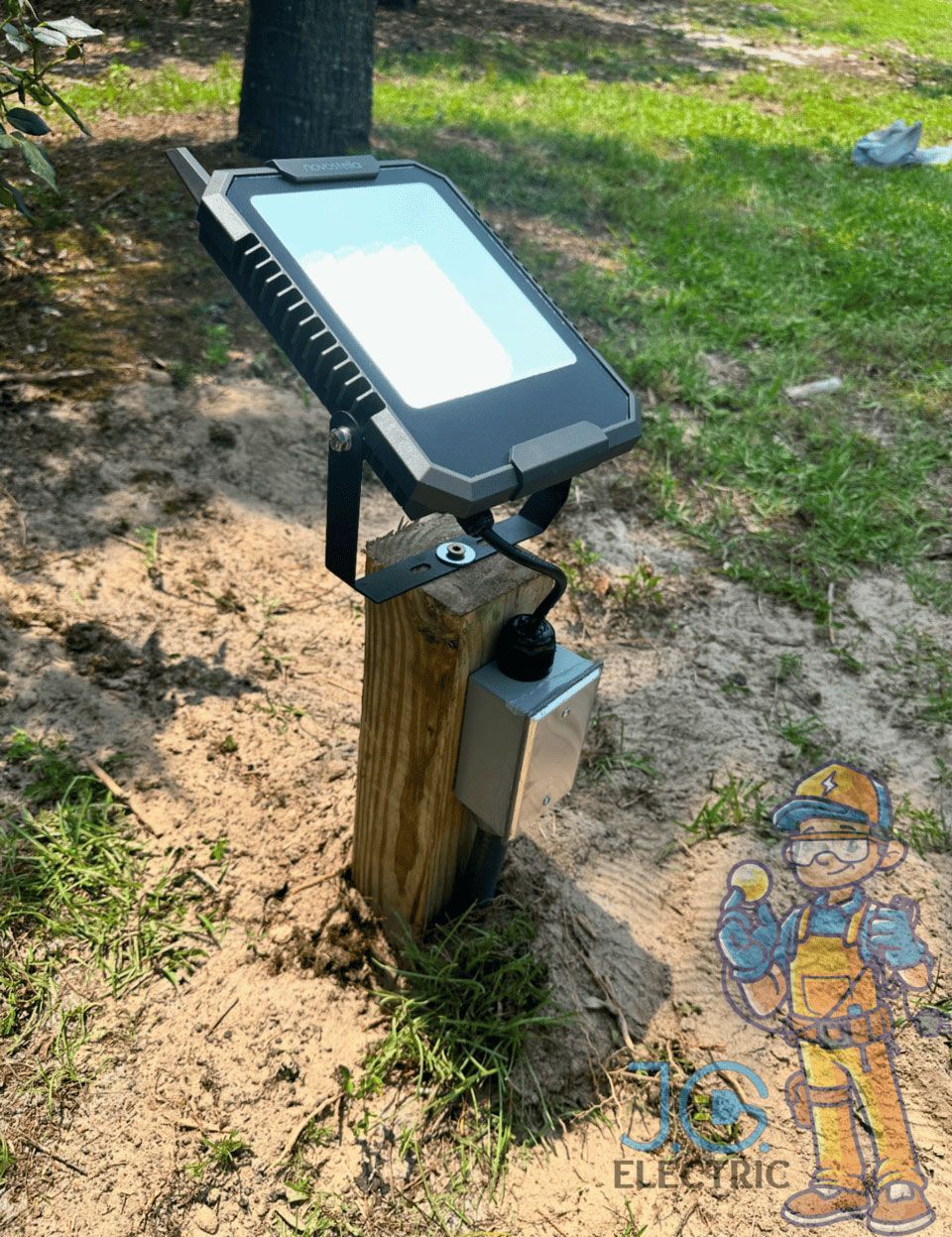 A small, rectangular outdoor LED floodlight mounted on a wooden post in a grassy area, connected to a grey junction box.