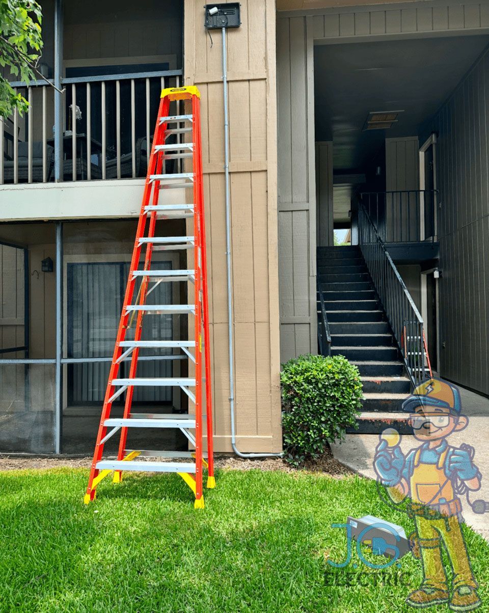 An orange fiberglass stepladder stands on grass next to a building wall with conduit, beside an outdoor staircase.