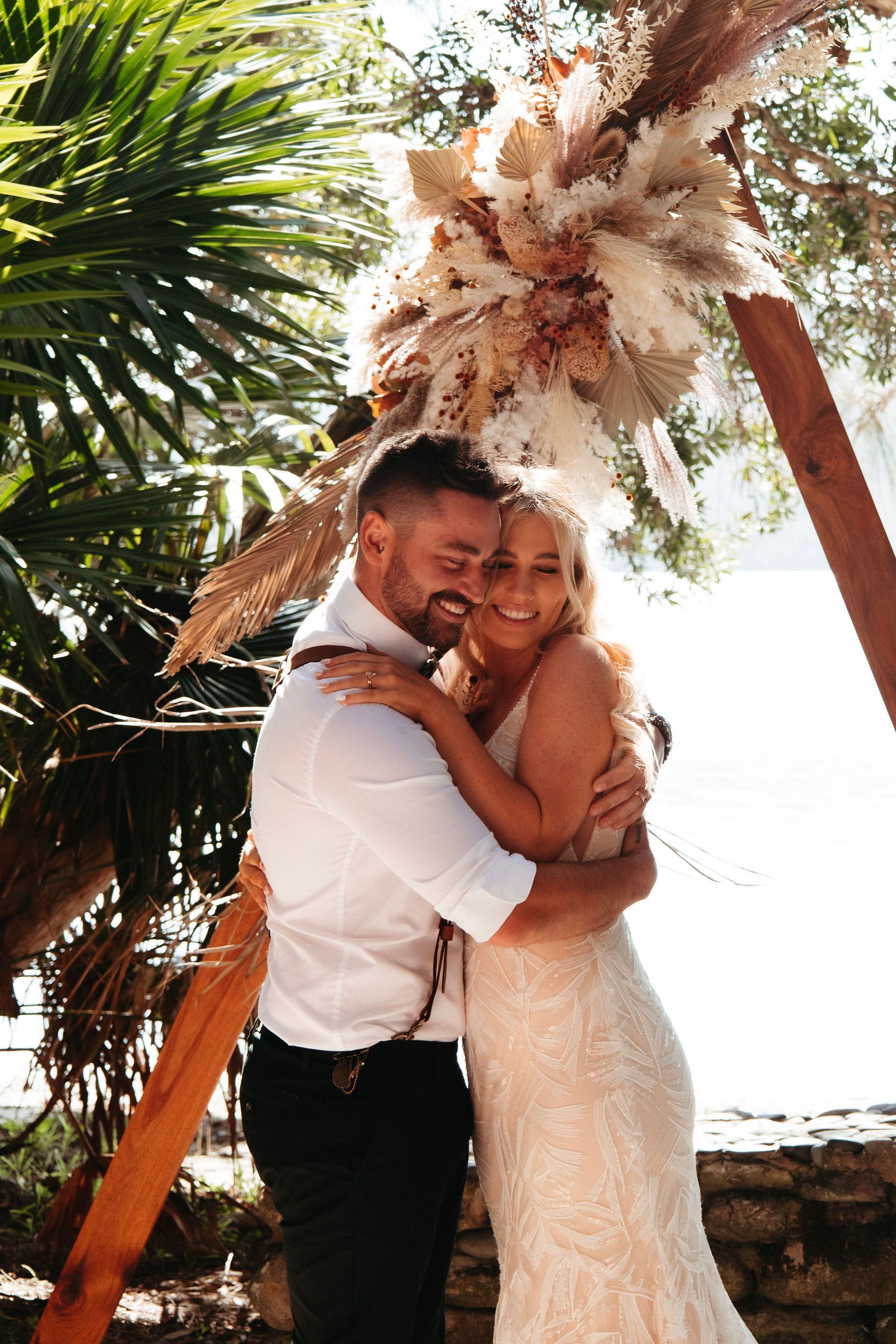 Bride and groom on beach, she in lace gown, he in suspenders, looking down, sunny day.