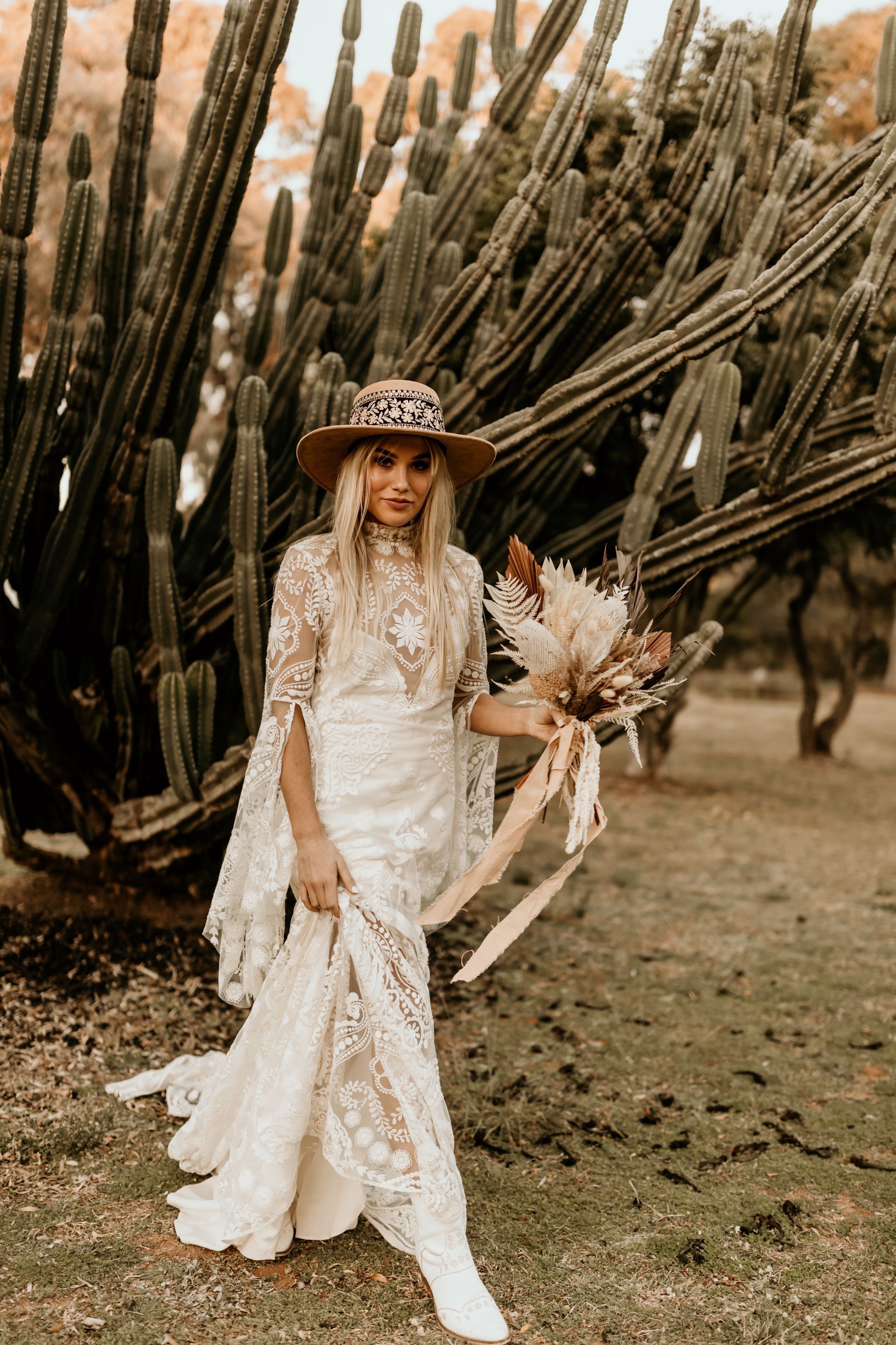 Bride in low-back dress carries bouquet, smiles at camera, walks beside groom on beach, sunlight.