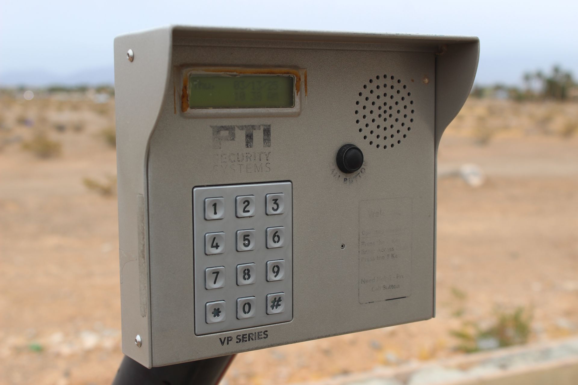 Gray keypad security system with display, speaker, and visor in a desert environment.