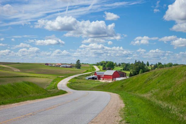 Winding rural road through green hills toward a red barn under a blue, cloud-filled sky