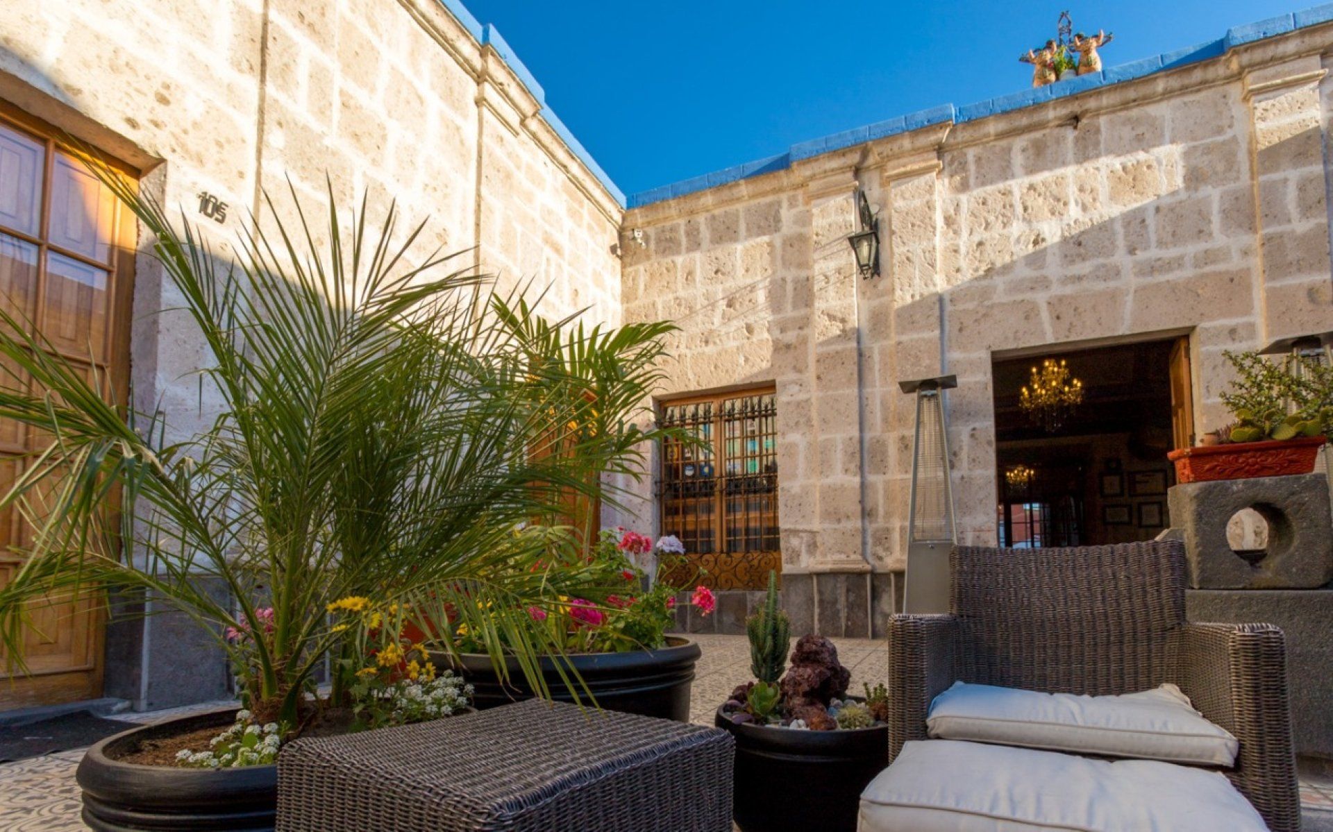 Sunny stone courtyard with wicker chairs, potted palms, and a metal door under a blue sky
