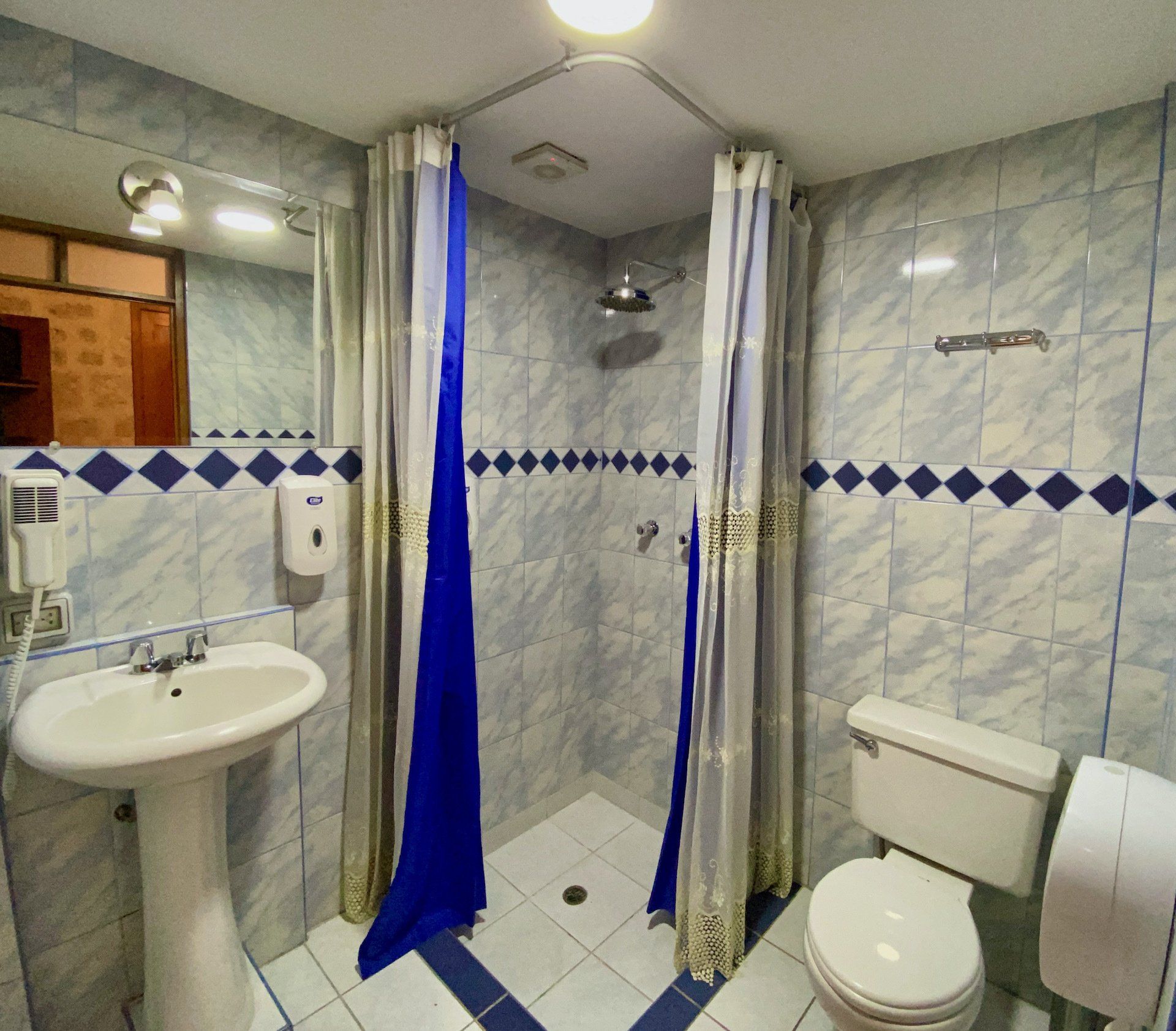 Compact bathroom with sink, toilet, shower stall, and blue curtain divider, tiled in white and blue.