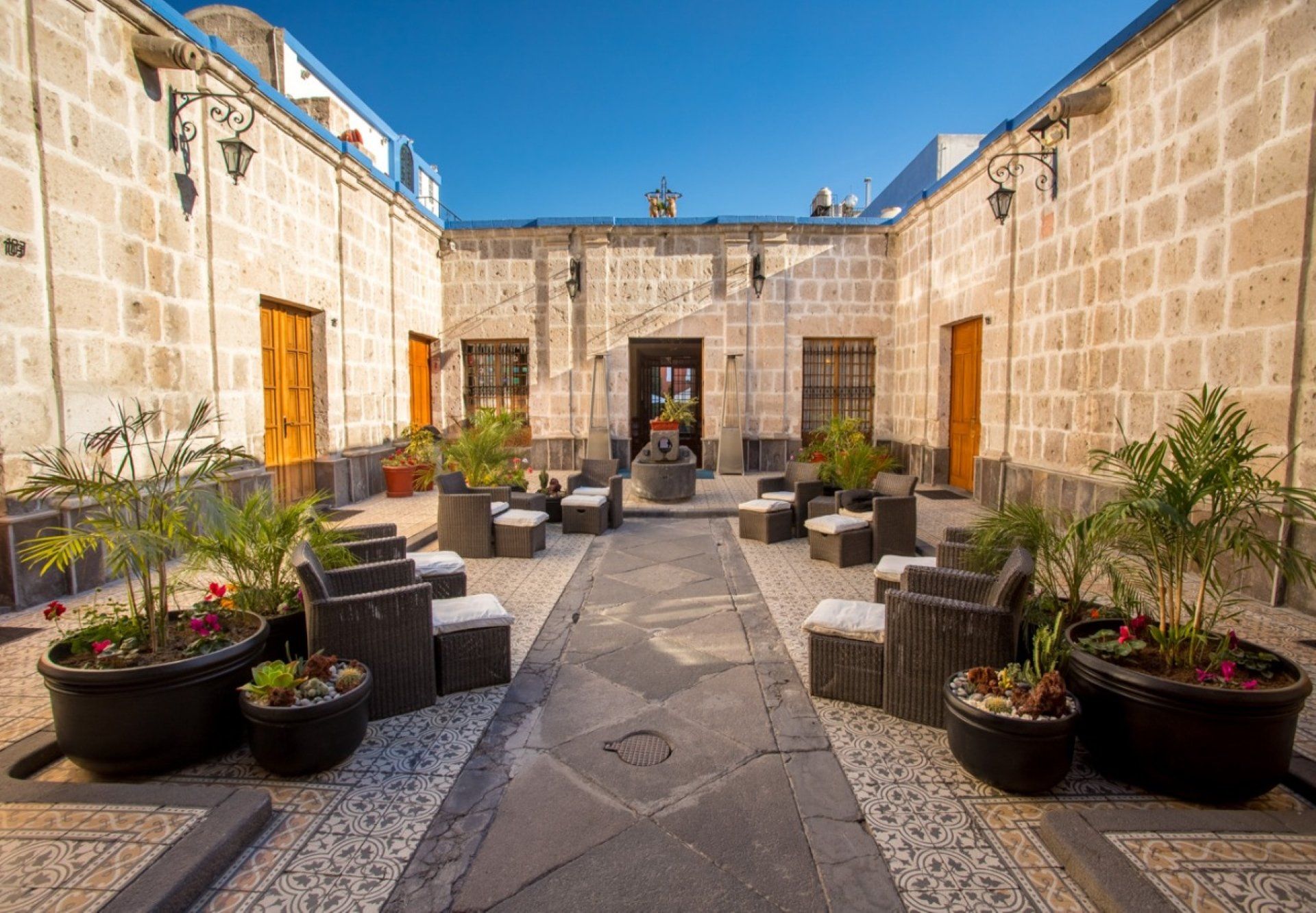 Stone courtyard with potted plants, black chairs, and tables under a bright blue sky