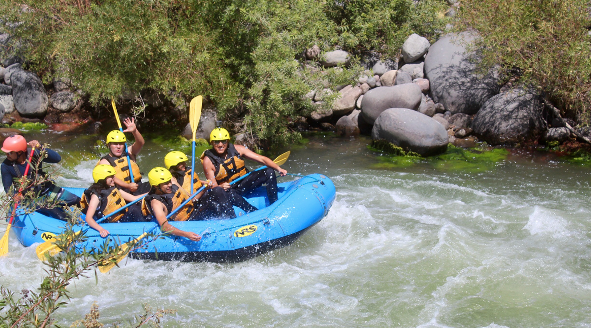 Personas haciendo rafting en una balsa azul, chapoteando en los rápidos. Llevan casco y remos.