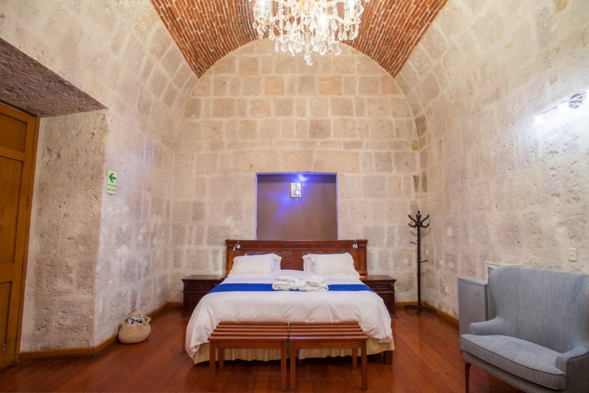 Stone-walled bedroom with arched ceiling, chandelier, wooden bed, and gray armchair