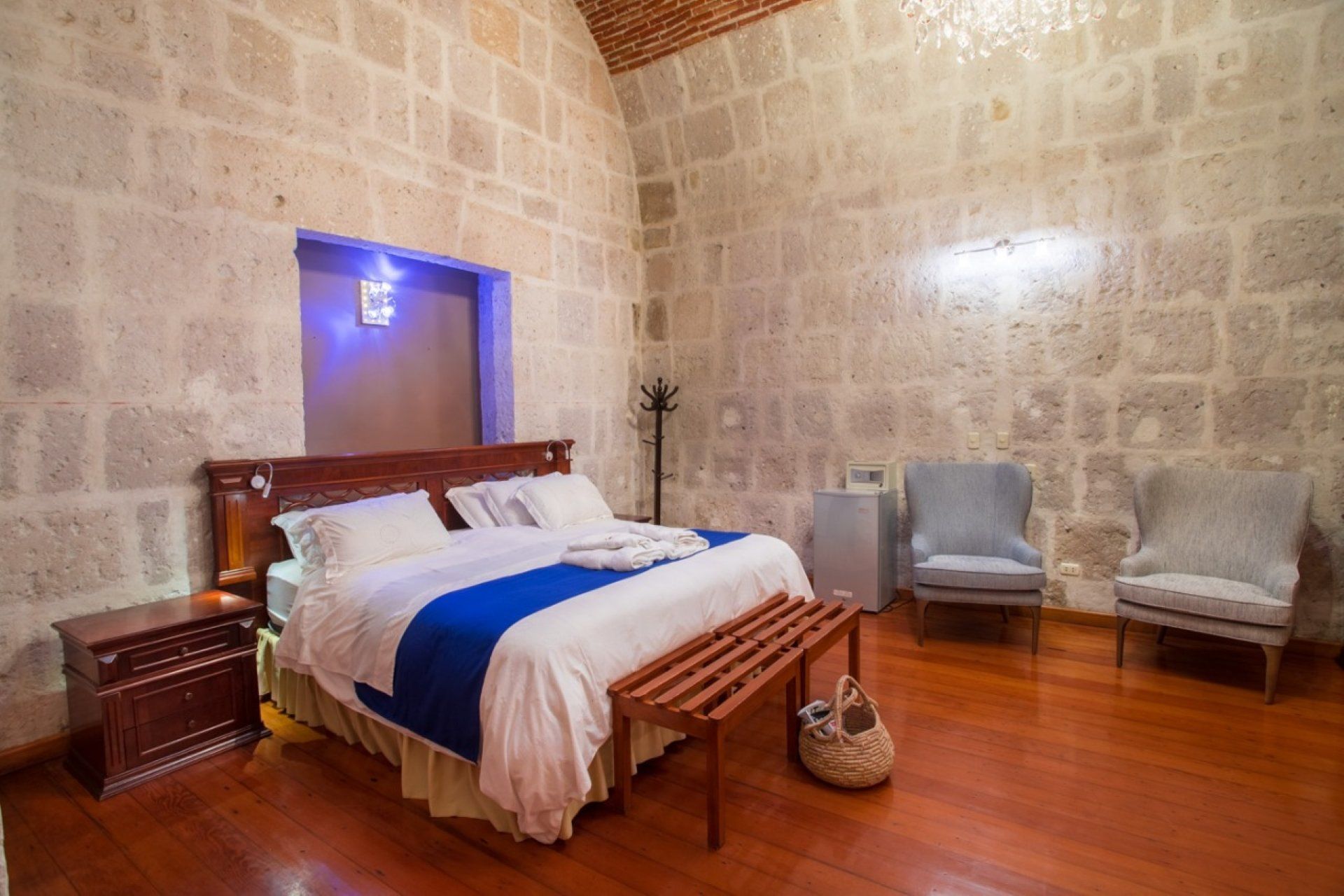 Cozy stone bedroom with a wooden bed, blue and white bedding, two chairs, and a guitar on the floor.