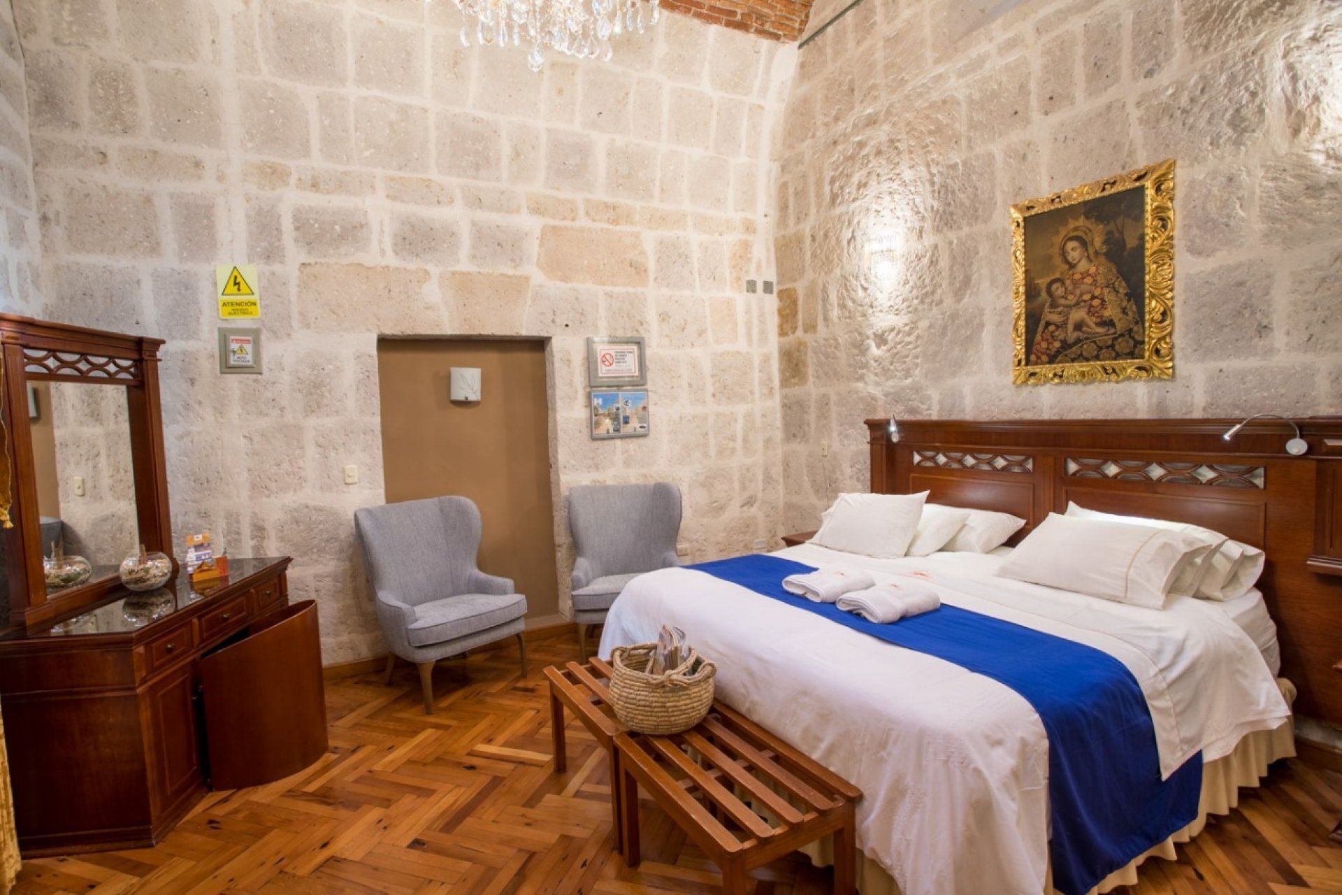 Stone-walled bedroom with a large bed, blue runner, chairs, and wooden furniture.