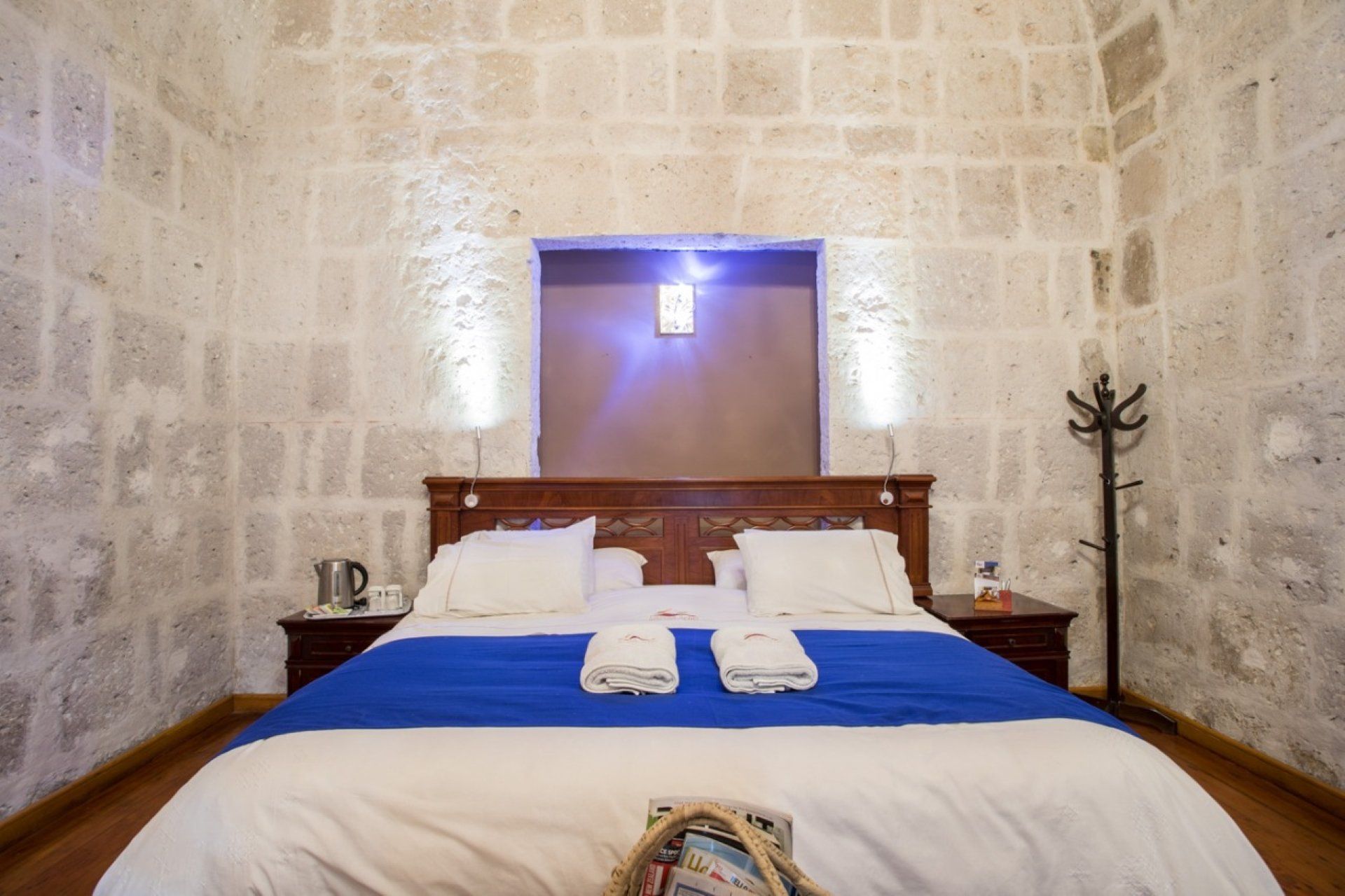 Stone-walled bedroom with a wooden bed, blue coverlet, white pillows, and a lit wall sconce above the headboard