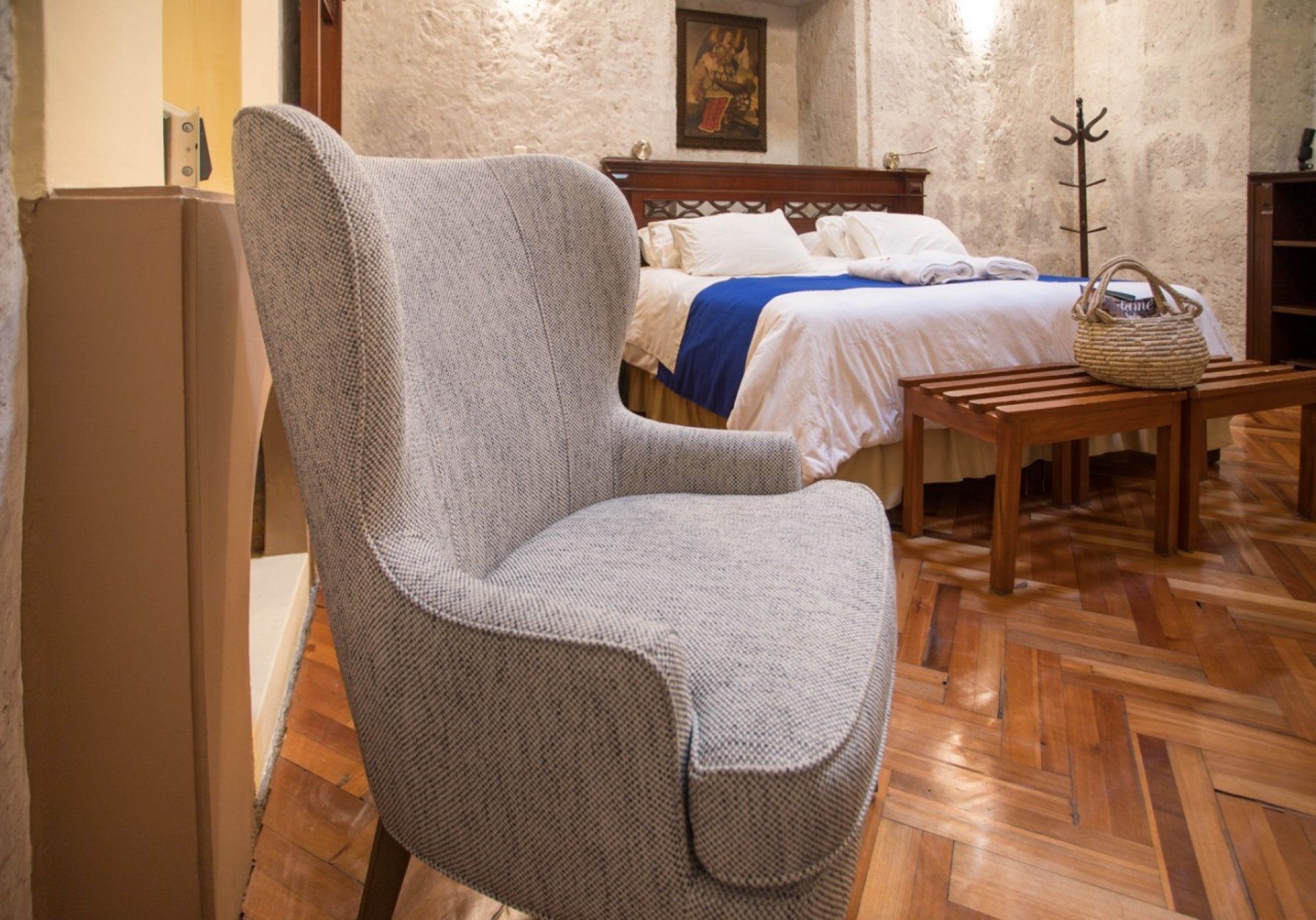 Cozy bedroom with a gray armchair in the foreground, bed with blue runner, and wooden floorboards