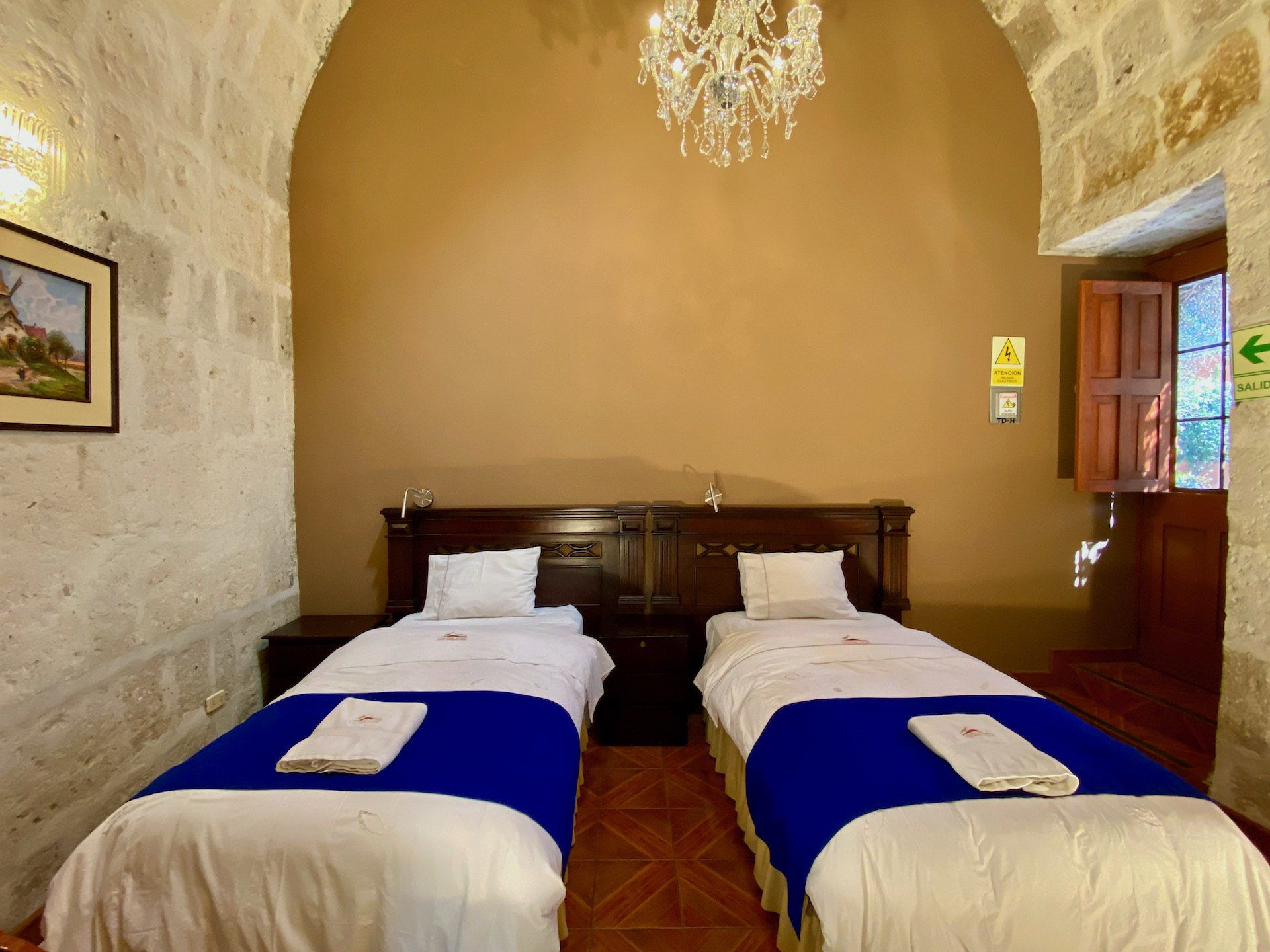 Two twin beds with blue runners in a stone-walled hotel room with an arched ceiling and chandelier