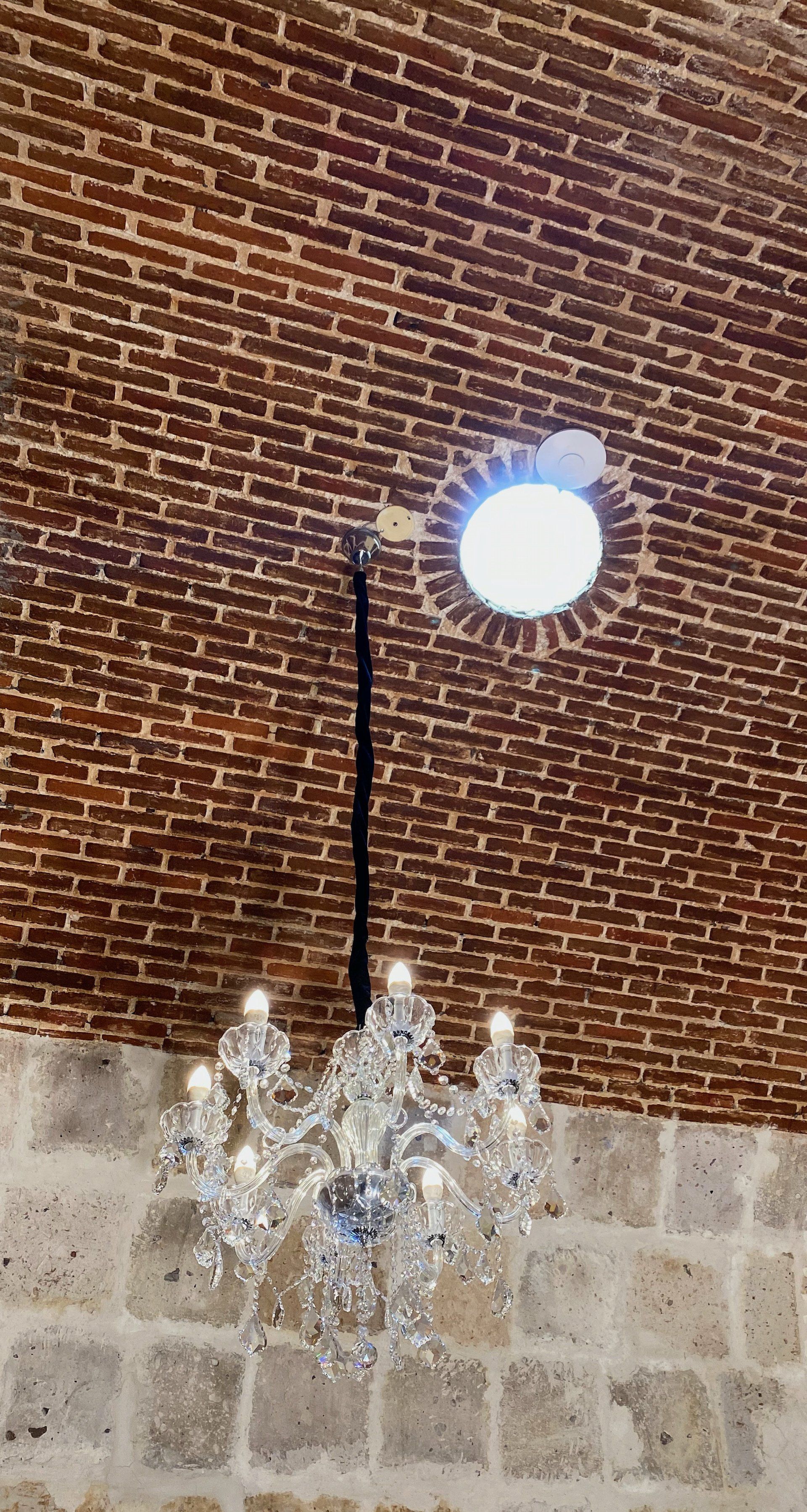 Chandelier hanging beneath a circular window in a brick and stone wall with bright daylight above