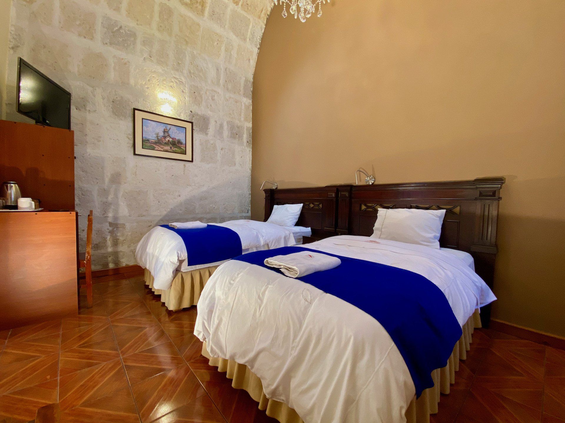 Hotel room with two twin beds, white bedding and blue runners, stone wall, and wooden floor.