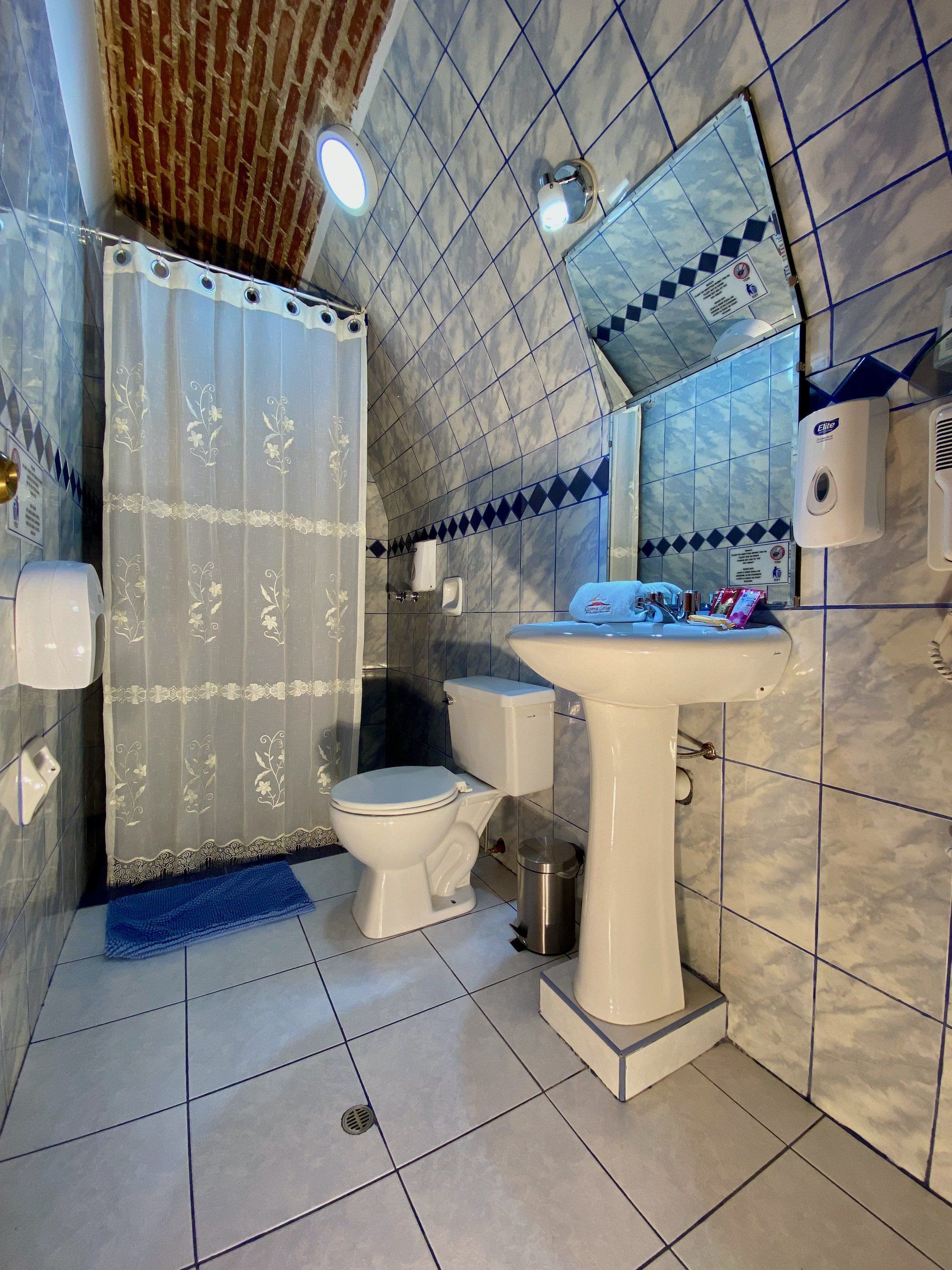 Small tiled bathroom with shower, toilet, and pedestal sink under bright lights.