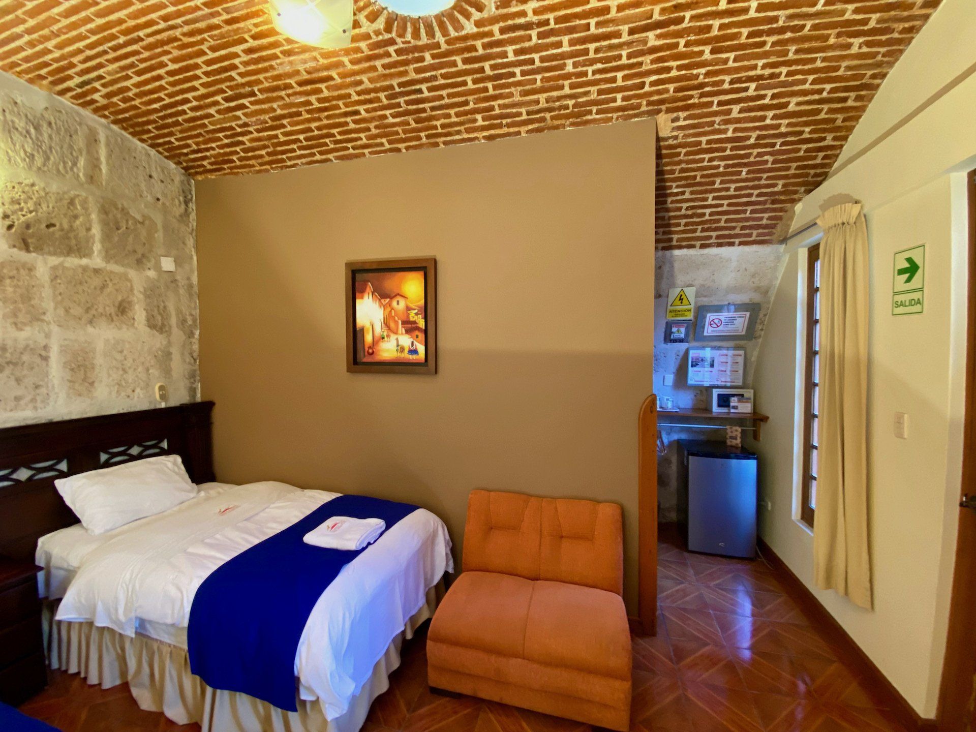 Small hotel room with a bed, orange chair, stone wall, framed art, and a mini-fridge area near the door