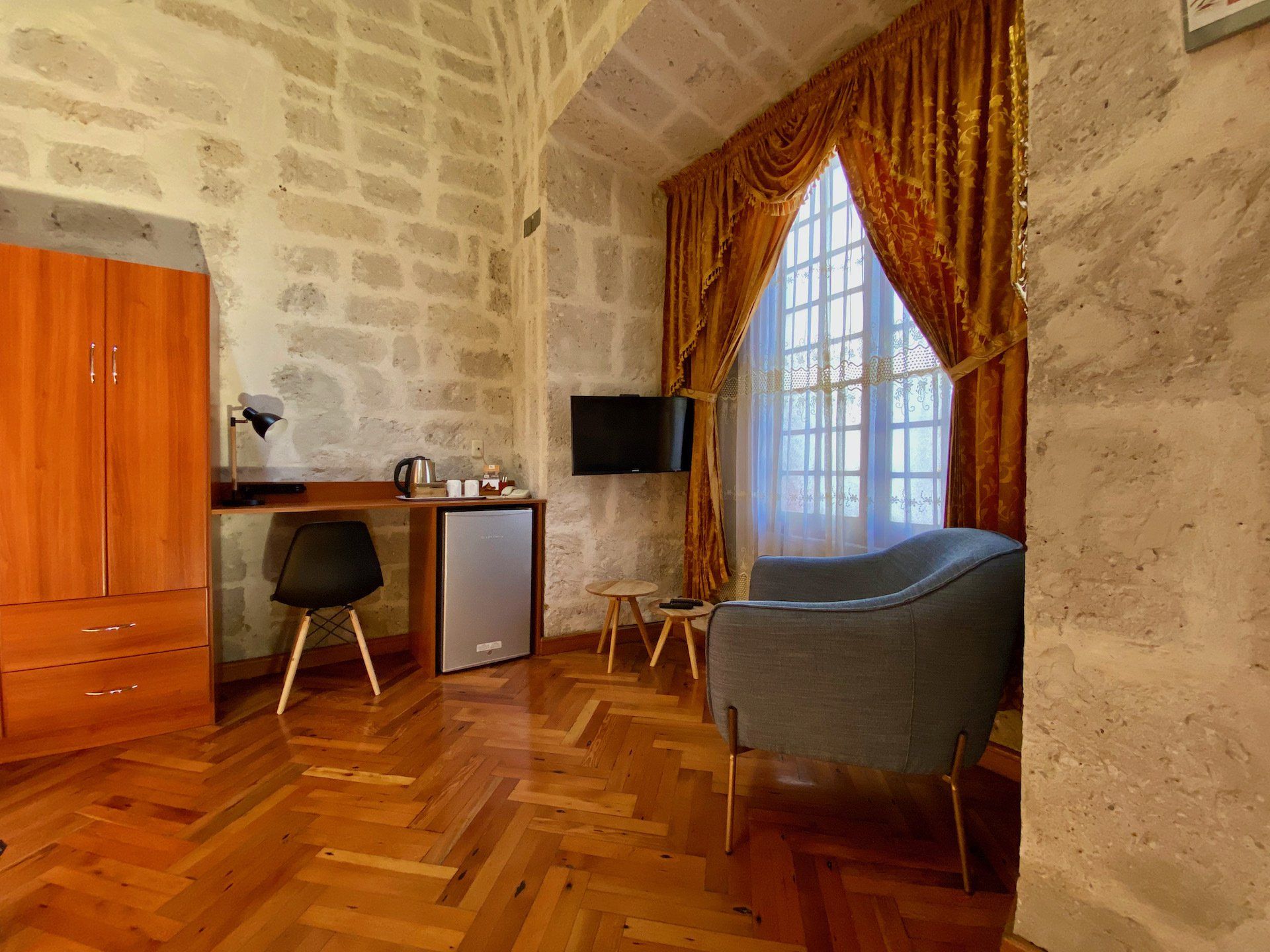 Stone-walled room with wooden floor, desk, chair, window with red curtains, and gray armchair