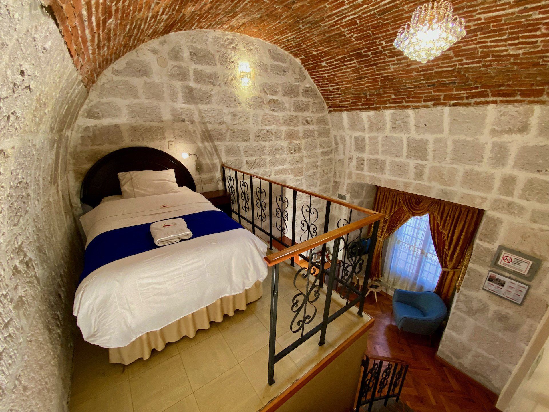 Stone-walled bedroom with arched ceiling, white bed, blue runner, and wrought-iron stair railing