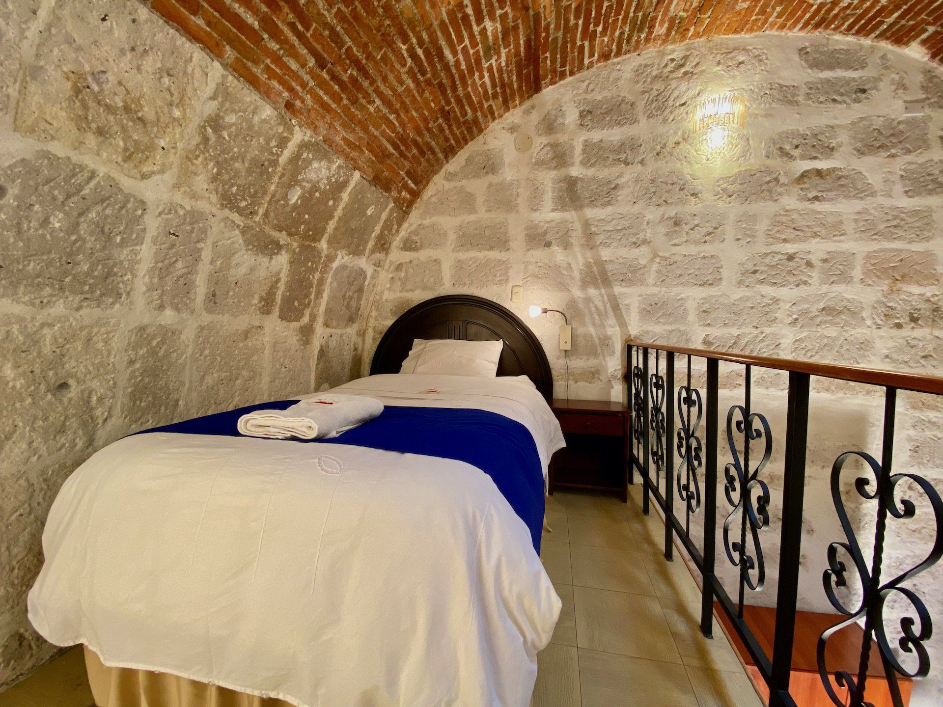 Stone-vaulted bedroom with a bed, white linens, blue blanket, and wrought-iron railing