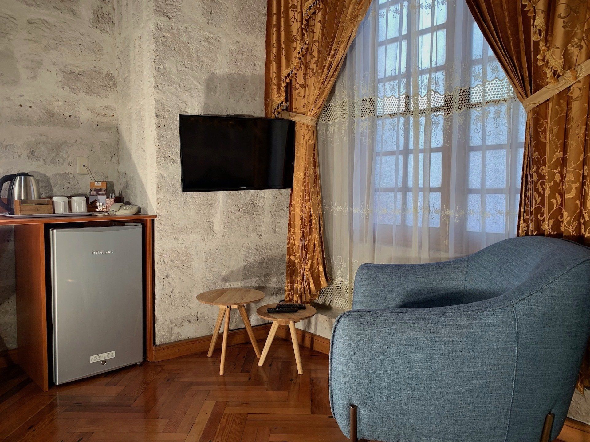 Cozy living room with blue armchair, wall-mounted TV, small fridge, and window with gold curtains