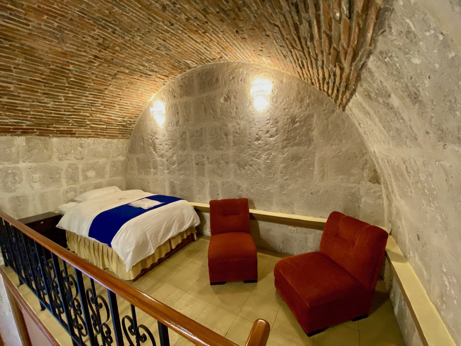 Stone-walled bedroom with brick vaulted ceiling, bed with blue blanket, and two red chairs