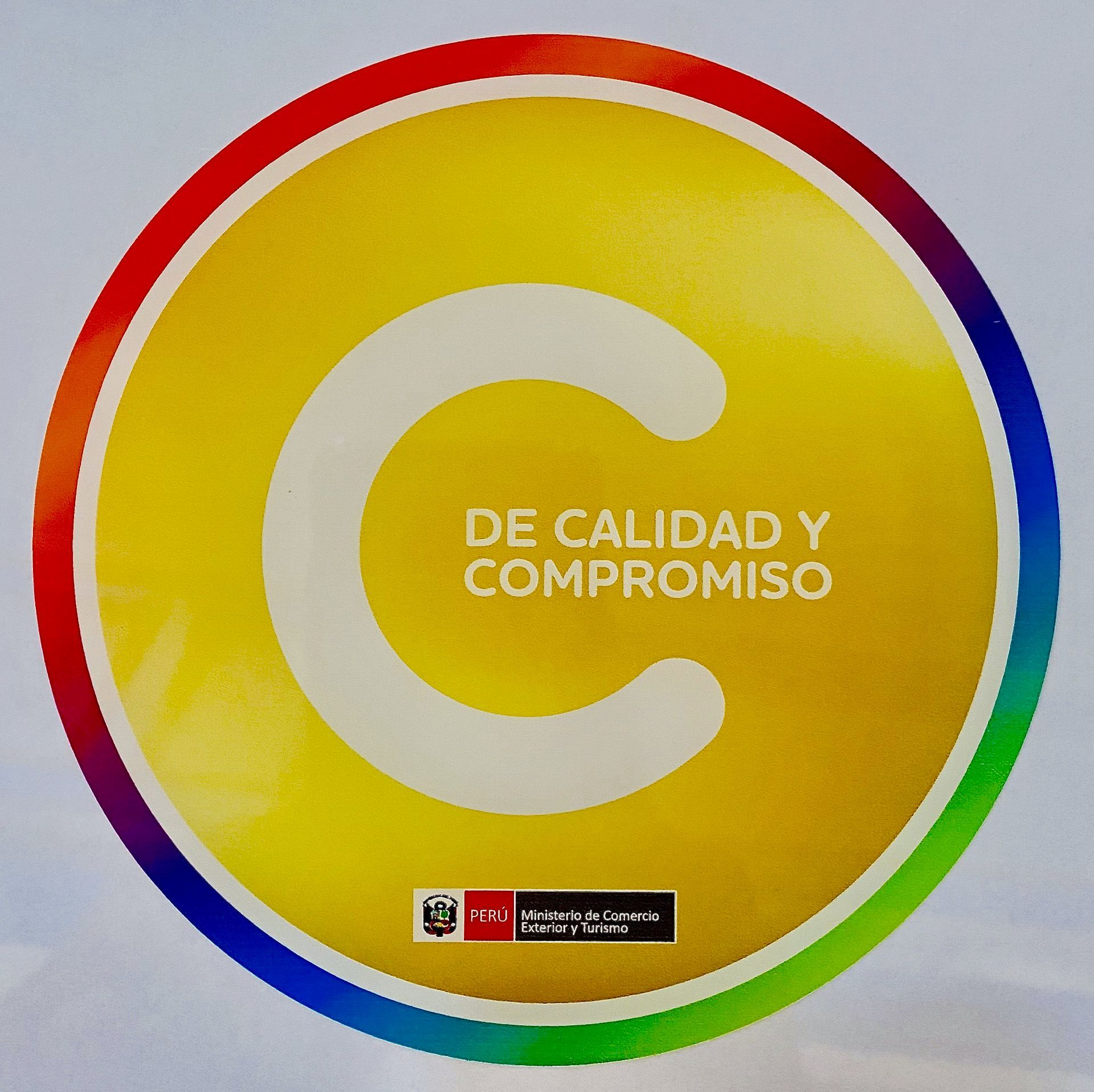 Circular yellow logo with multicolor ring and the words “DE CALIDAD Y COMPROMISO” in the center.