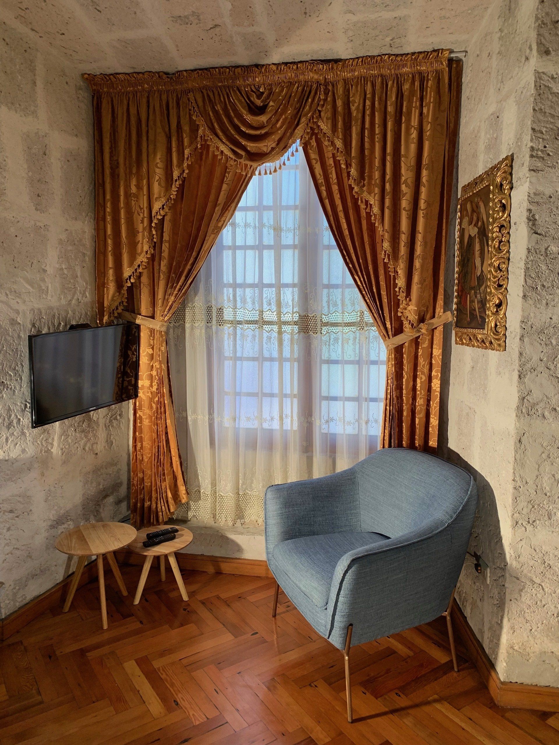 Cozy corner with blue chair, small stools, wall-mounted TV, and gold drapes over a window