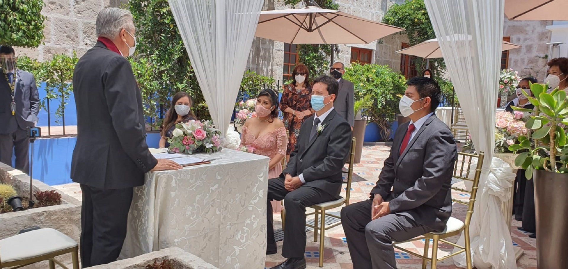 Man speaking to seated guests at an outdoor event under a white canopy, with flowers and potted plants nearby