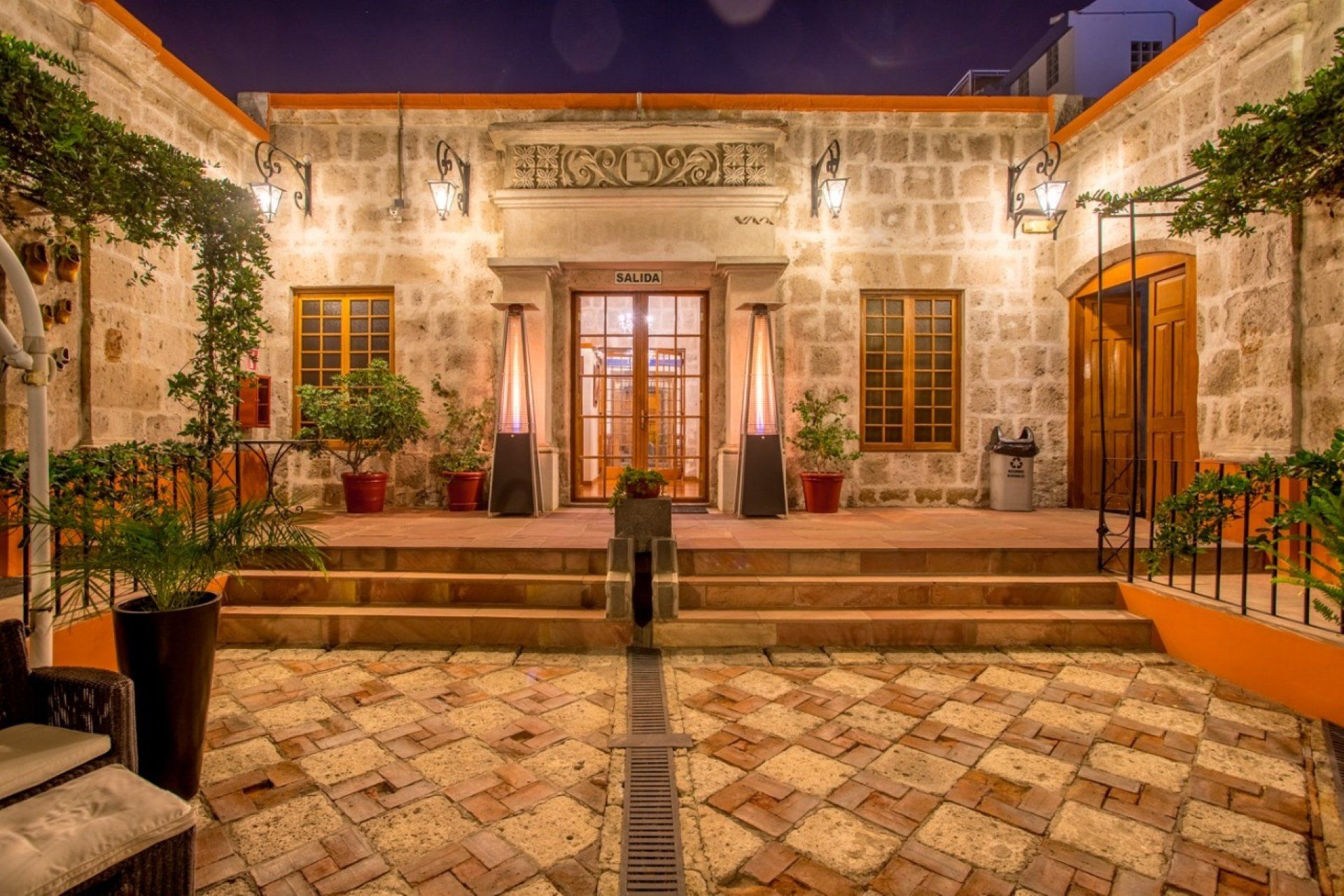 Illuminated courtyard facade with stone steps, central wooden door, and potted plants at dusk