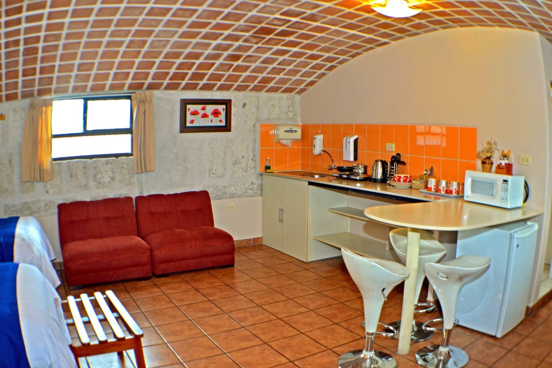 Small modern kitchen with red sofa, orange cabinets, tiled floor, and round glass table with white chairs