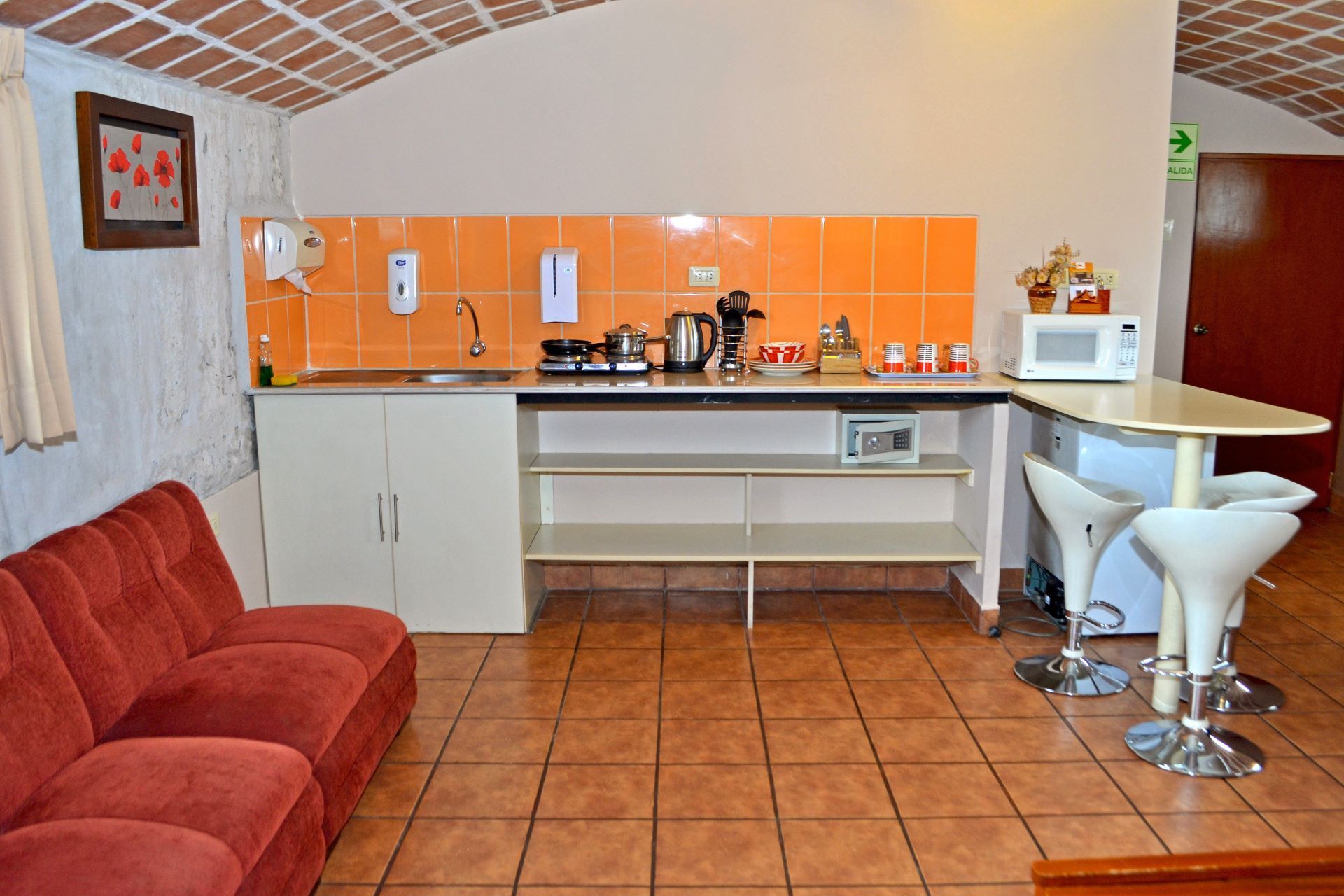 Compact kitchen with orange tile backsplash, white cabinets, red sofa, and bar stools on a tiled floor