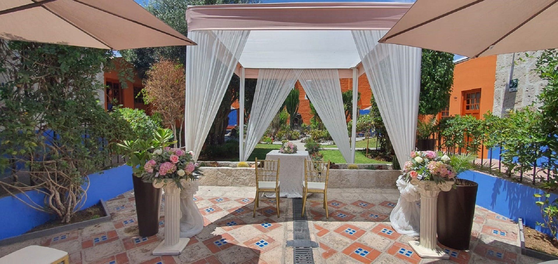 Elegant outdoor wedding canopy with white drapes, floral accents, and two chairs on a tiled patio garden.
