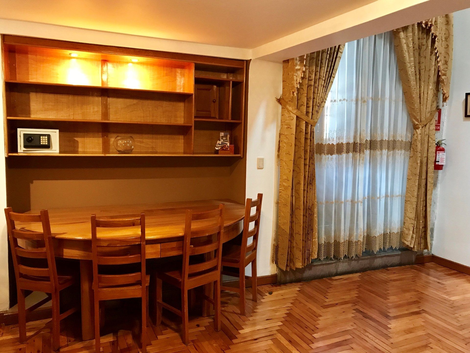 Dining room with wooden table and chairs, built-in cabinet, and sheer curtains over a window.