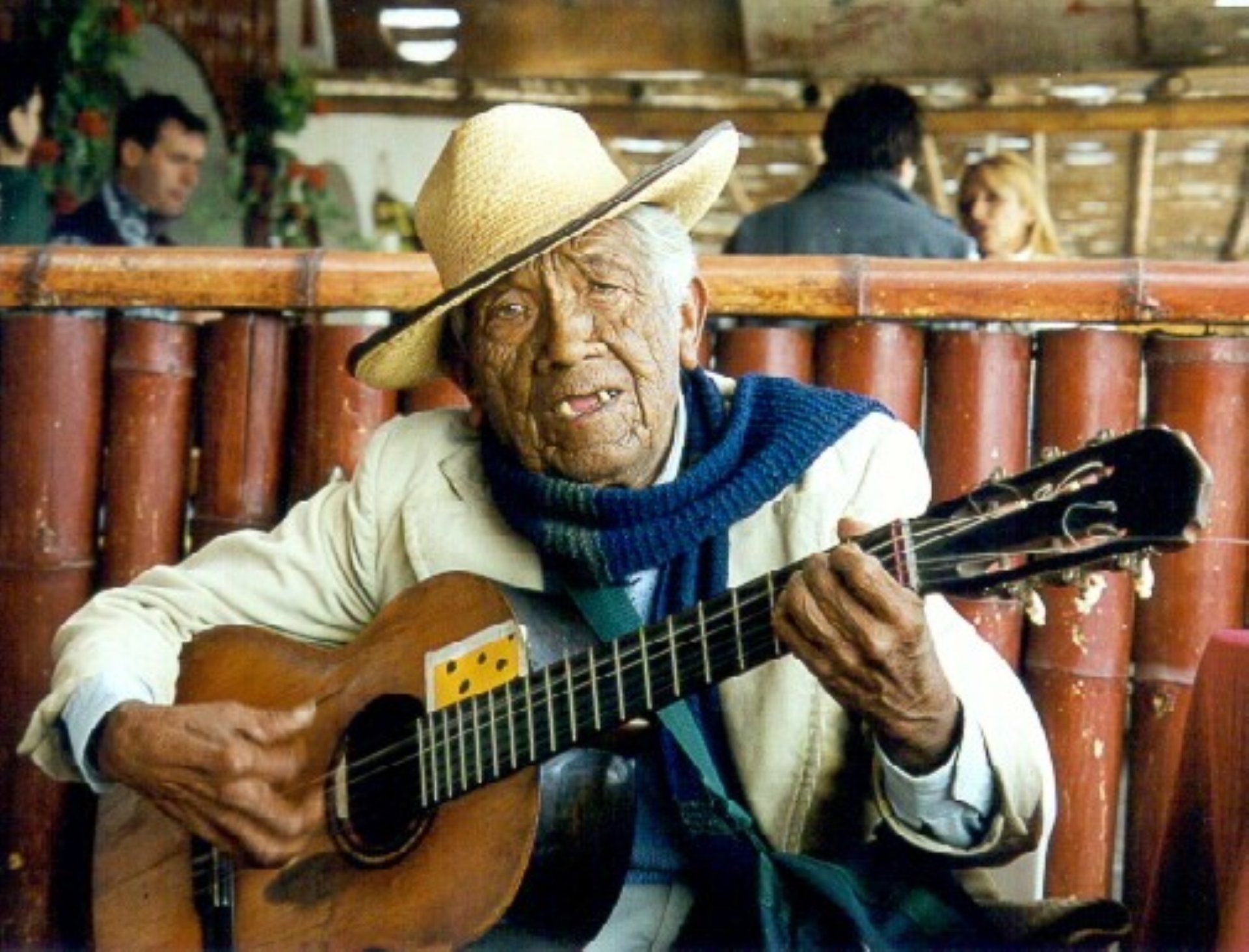 Man in straw hat playing guitar indoors, seated among bamboo chairs, with a relaxed smile.