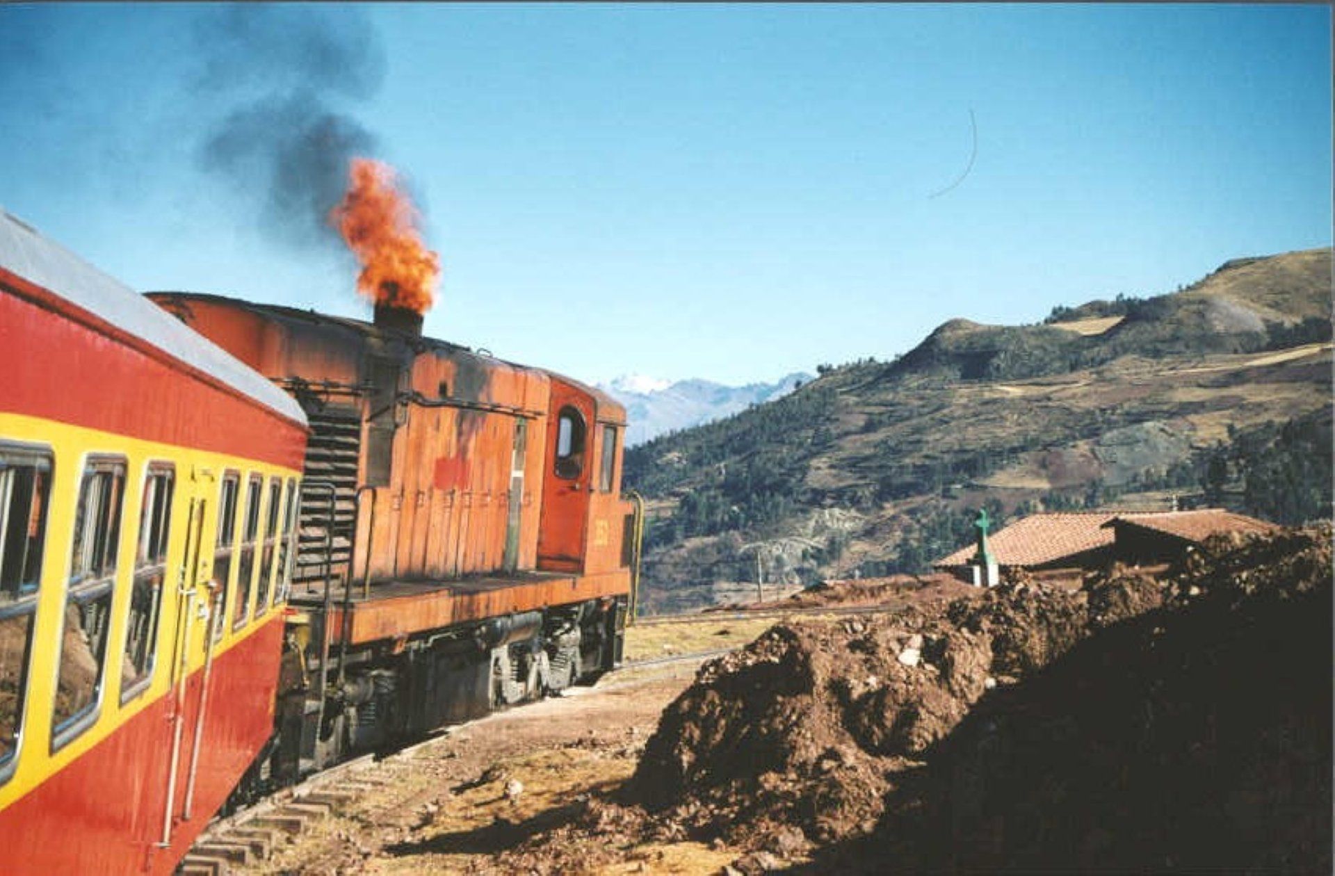 Orange locomotive pulling a red-and-yellow train through a mountain landscape with smoke rising.