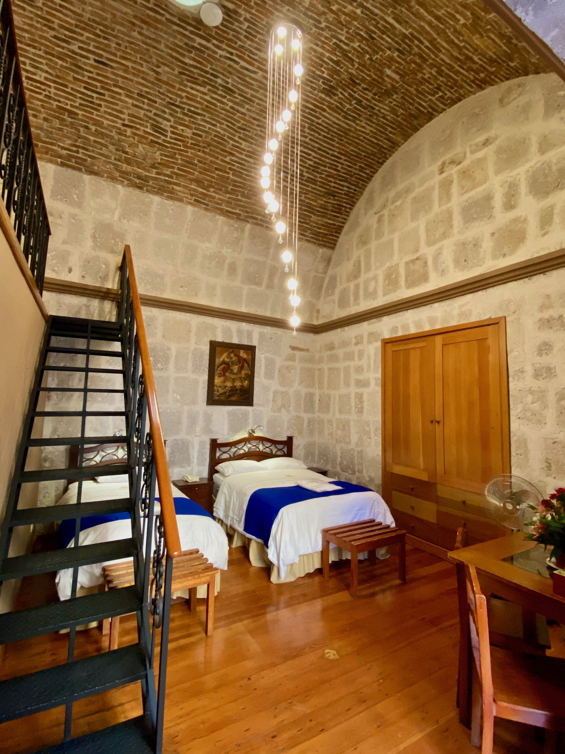 Stone-walled bedroom with a single bed, wooden door, loft stairs, and arched brick ceiling
